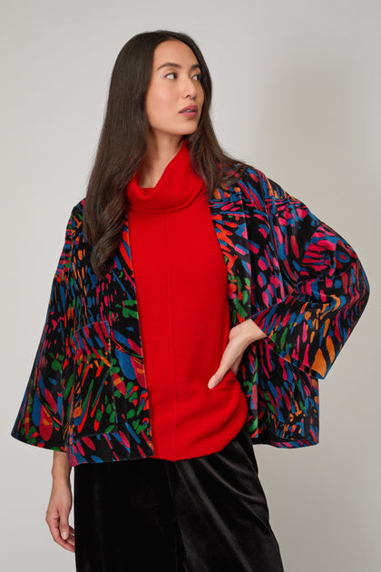 A woman wearing the Skyfire Velvet Velvet Jacket over a red turtleneck sweater and black pants stands against a plain background, looking to the side.