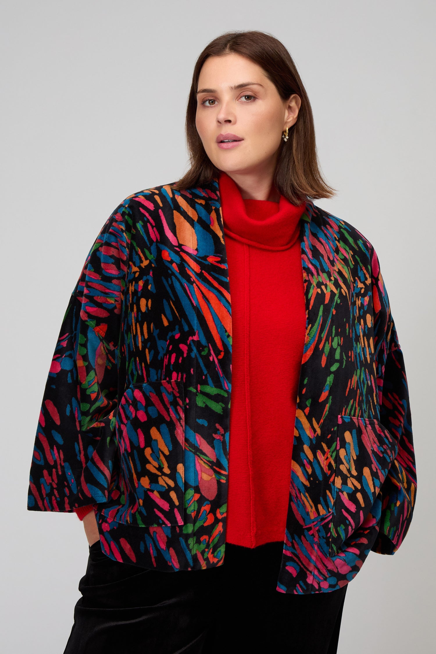 A woman wears the Skyfire Velvet Velvet Jacket, featuring colorful abstract patterns on black velvet over a red turtleneck, against a plain light background—a bold statement piece for chic evening style.