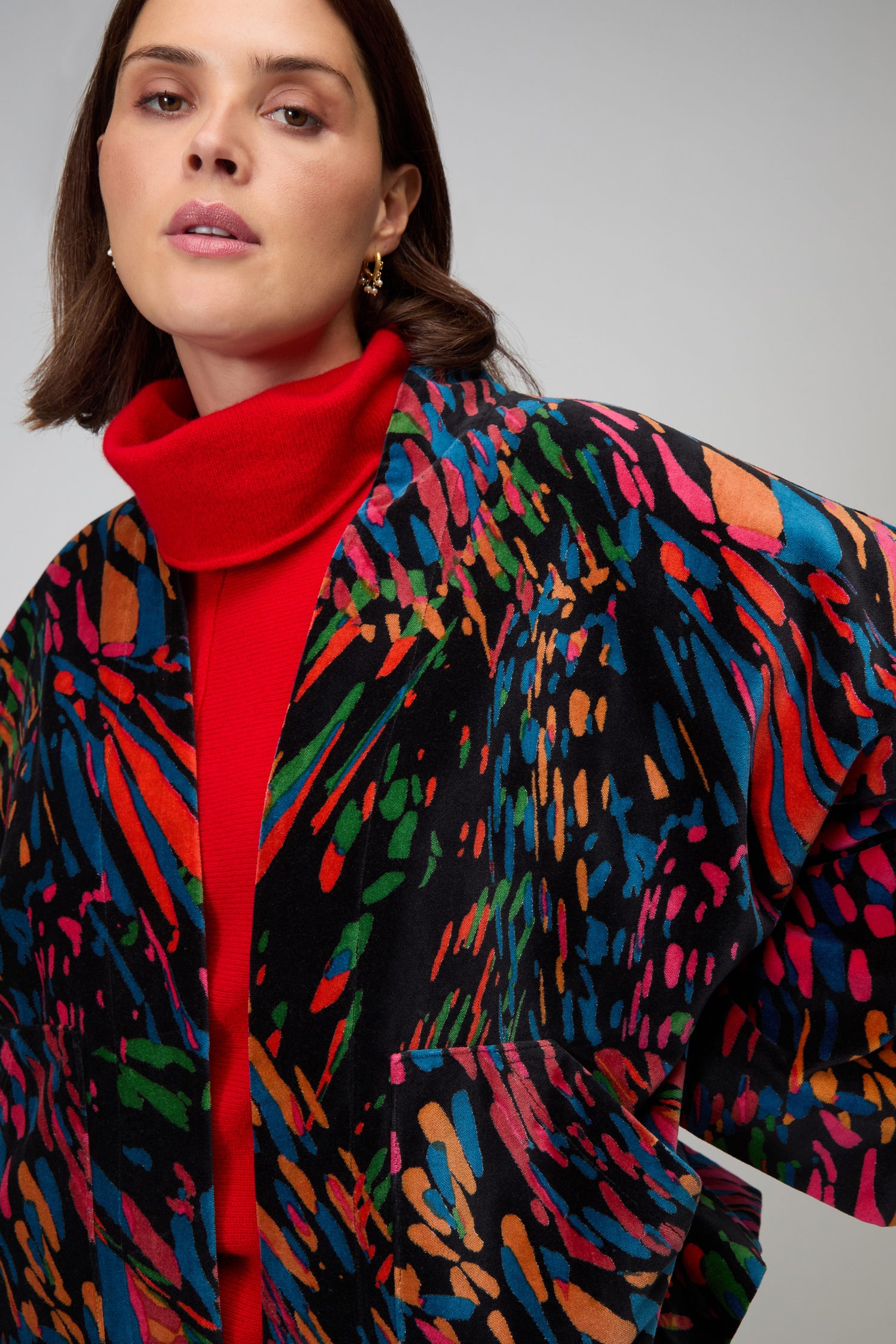 A woman models the Skyfire Velvet Jacket—a bold, multicolored statement piece—layered over a red turtleneck, standing against a plain gray background.