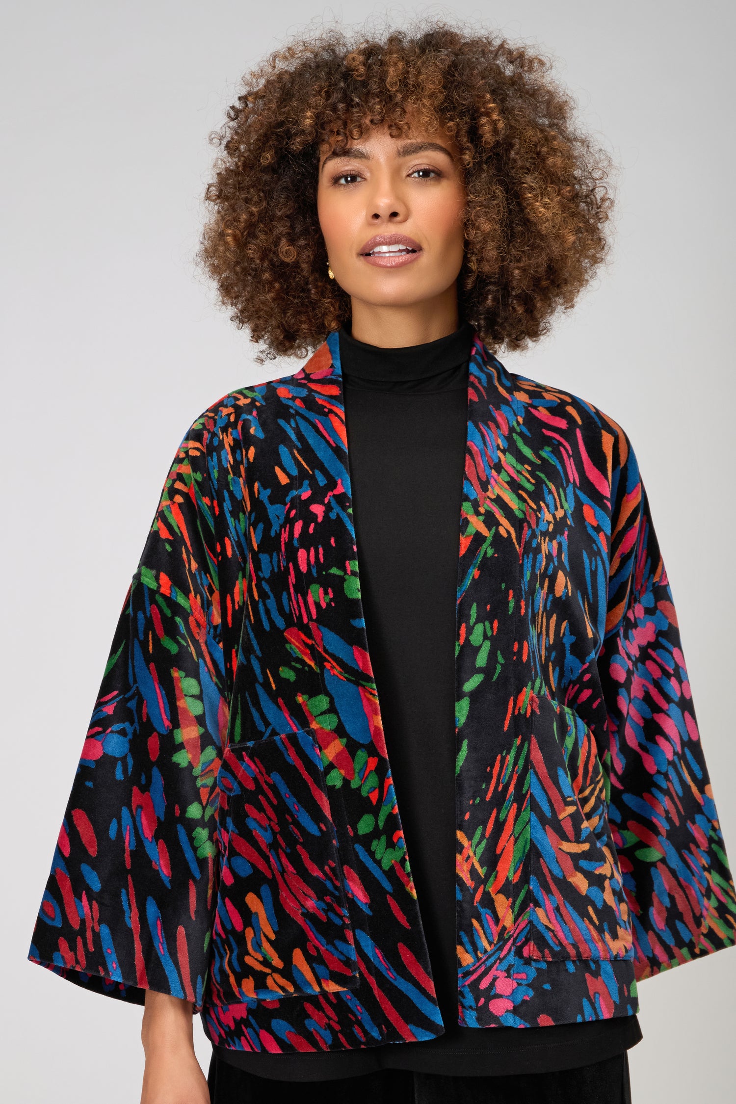 A woman stands against a plain light gray background, wearing the Skyfire Velvet Jacket—a colorful, patterned piece with wide sleeves and black turtleneck—perfect as a statement layer for chic evening looks.