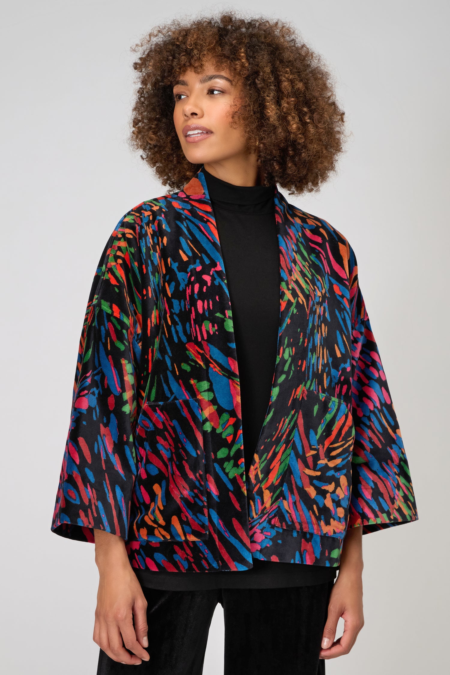 A woman stands against a plain light background, wearing a black turtleneck layered with the Skyfire Velvet Jacket—a colorful statement piece featuring wide sleeves. Ideal for elevating your evening look.