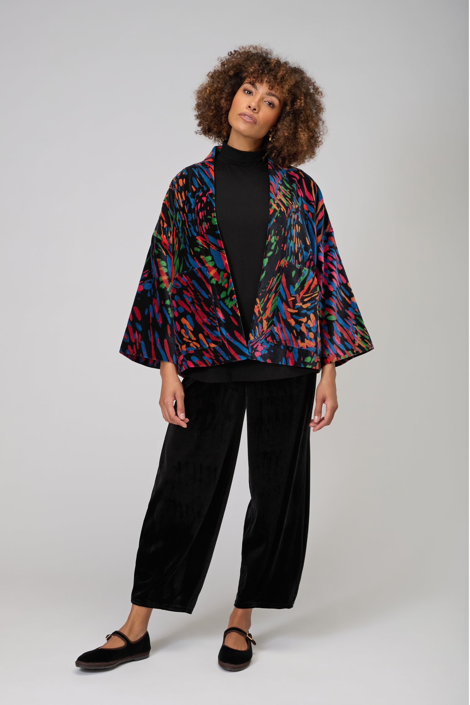 A woman stands against a plain background, wearing a black turtleneck, wide-leg black pants, black shoes, and the Skyfire Velvet Jacket as a statement layer for chic evening looks.