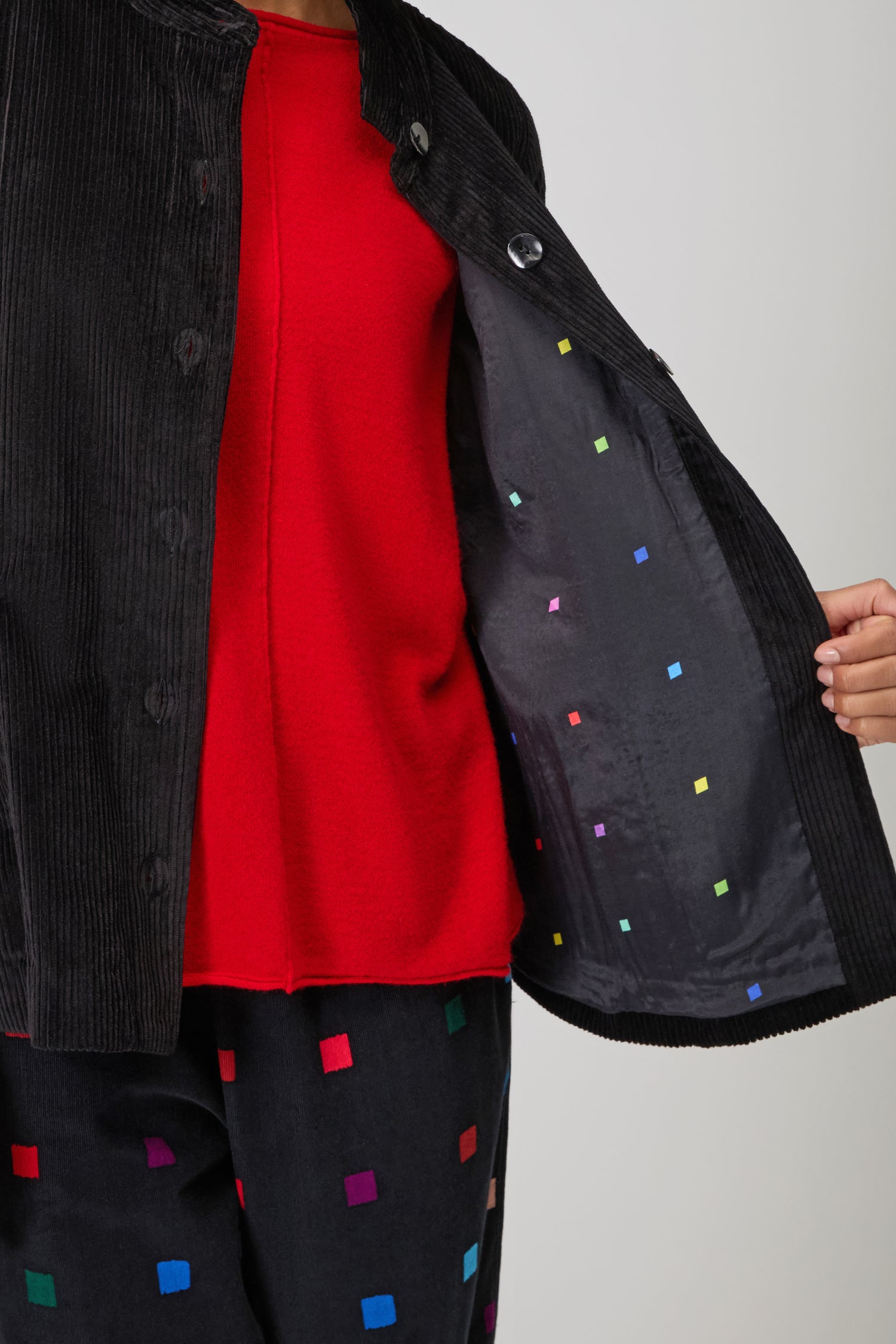 A person wears a red shirt with a black Jumbo Cord Jacket lined with small colorful squares, paired with matching black pants featuring similar colored squares.