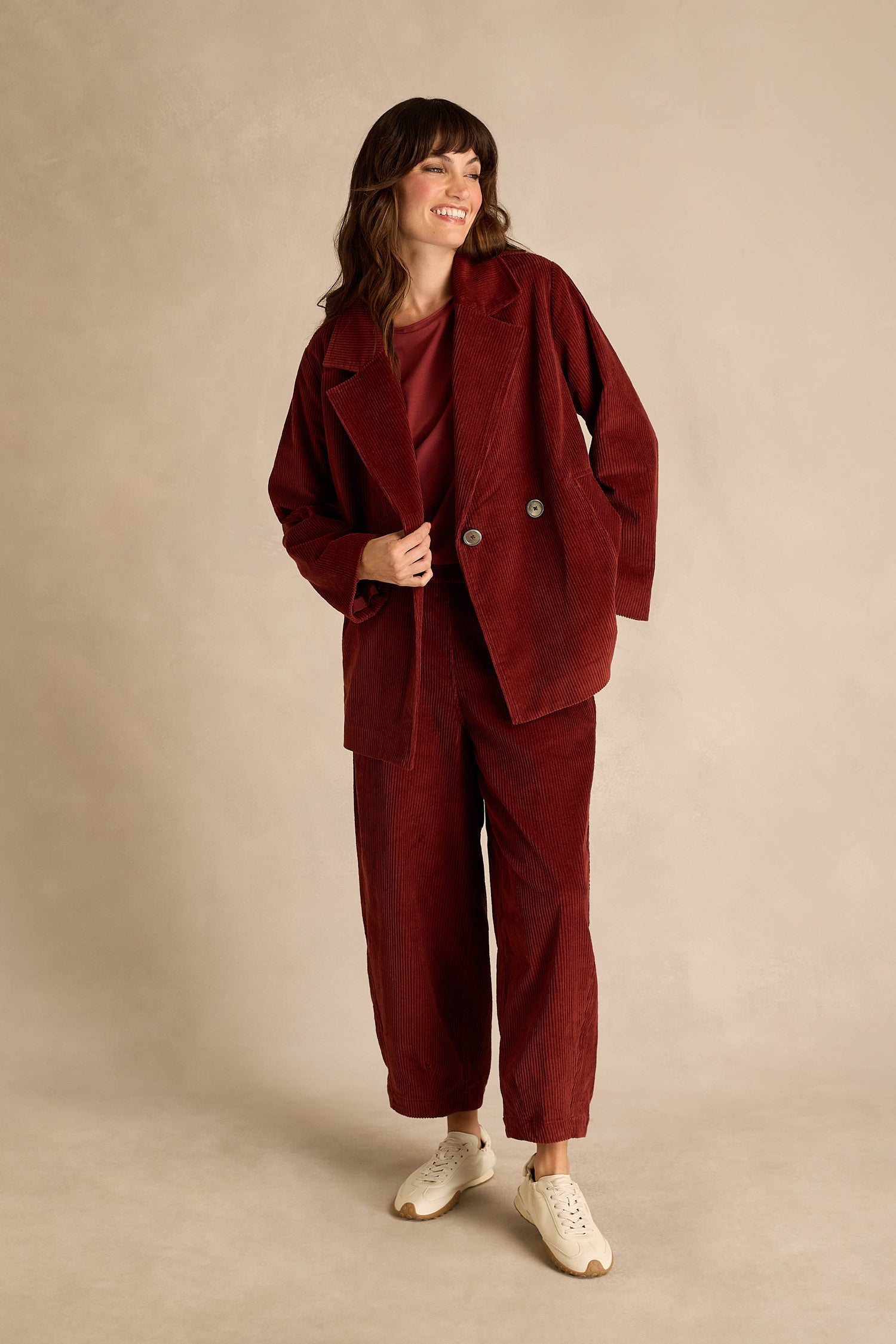 A woman stands smiling against a plain beige background, wearing the Conker Jumbo Cord Caban Jacket with matching maroon pants, a maroon top, and white sneakers.
