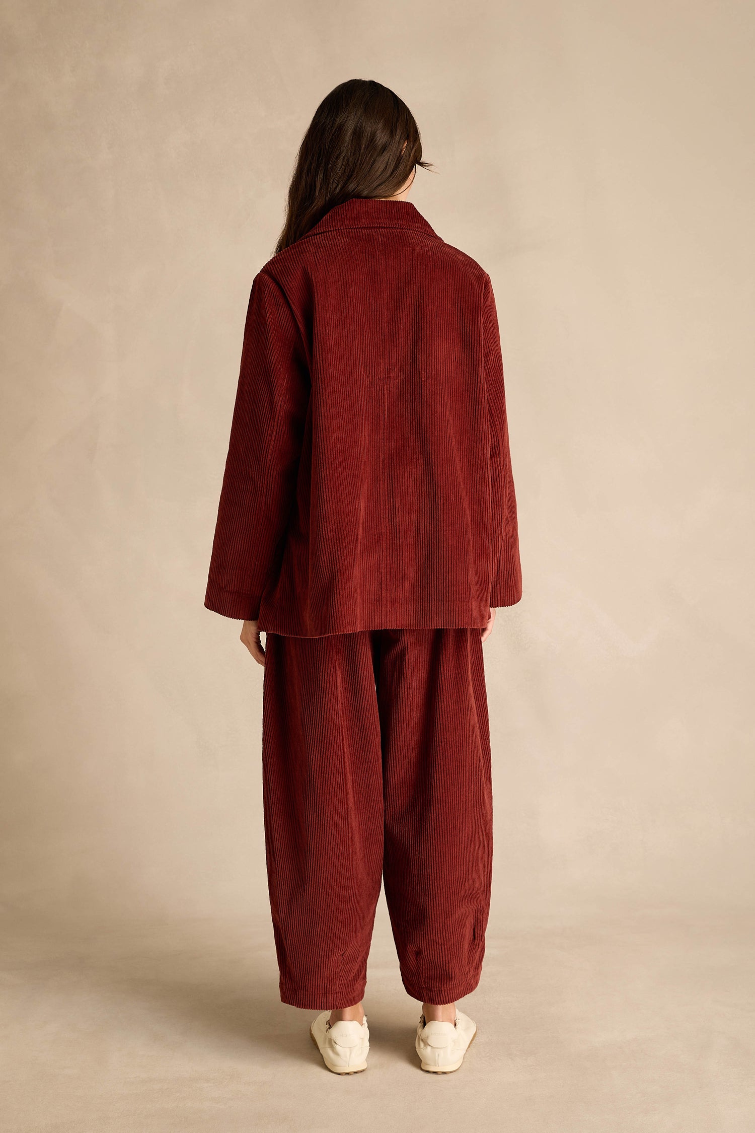 A person stands with their back to the camera, wearing the Conker Jumbo Cord Caban Jacket in a loose-fitting, dark red broad wale cord and white sneakers, set against a plain beige background.