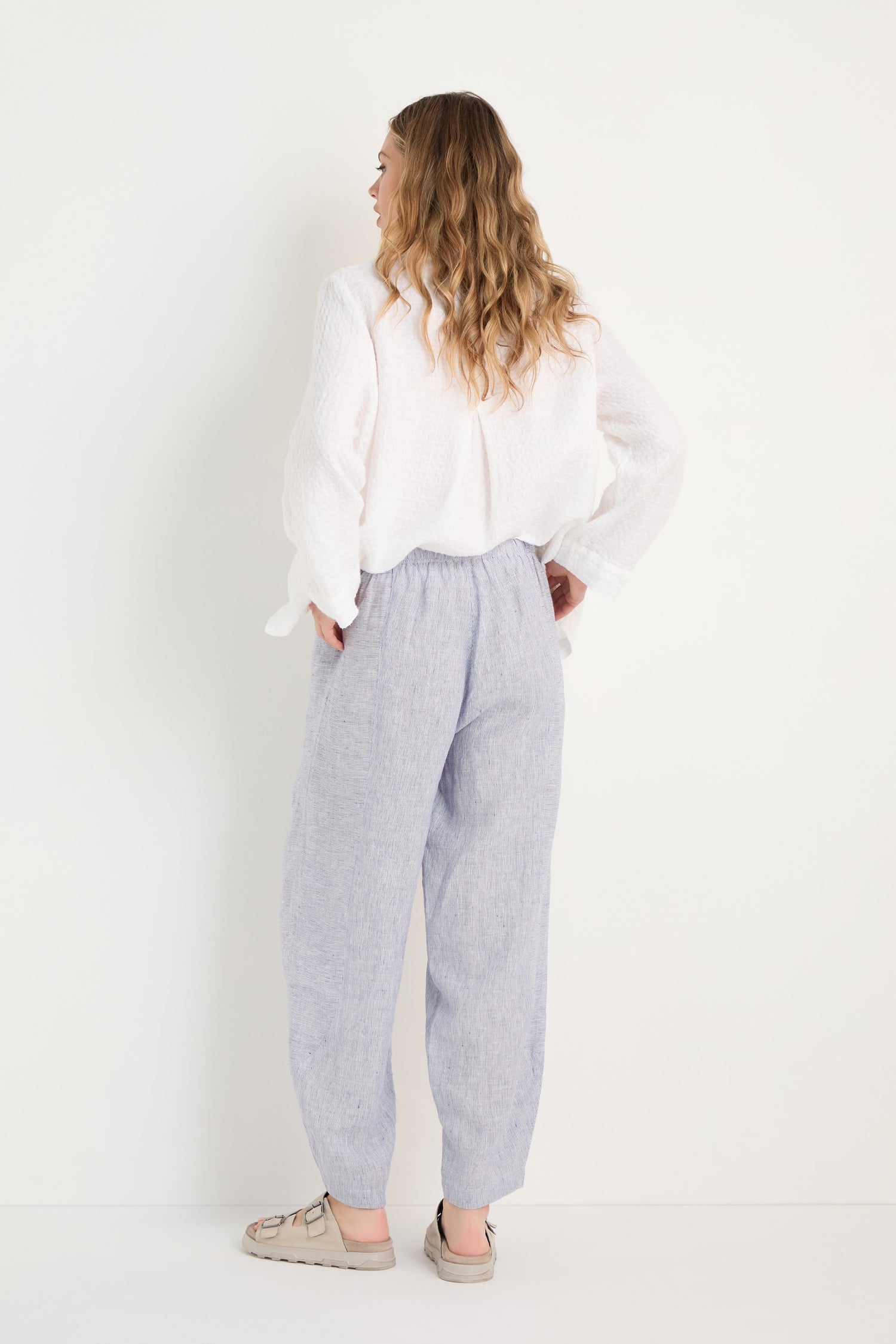 Woman stands against a white background, facing away, wearing a white textured blouse with Ticking Linen Stripe Bubble Trousers featuring an elasticated waist and beige sandals.