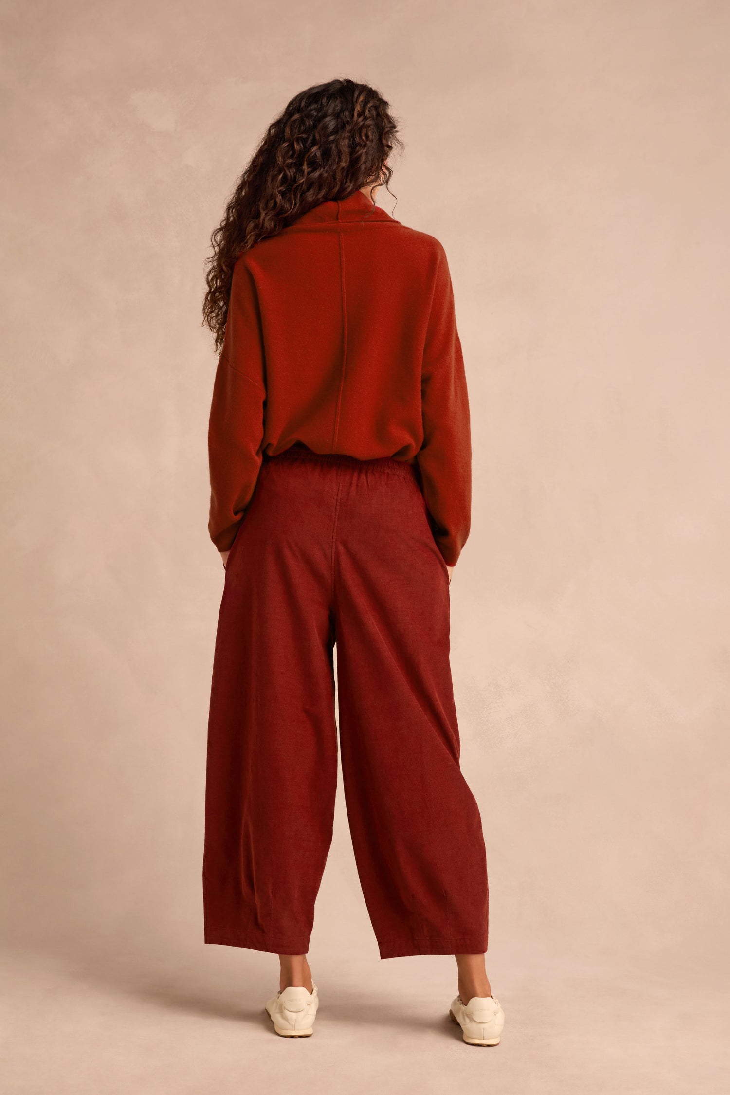 A person with long curly hair stands facing away, wearing relaxed-fit Conker Pinwale Cord Bubble Trousers and a rust-colored fine pincord sweater, paired with white shoes against a neutral background.