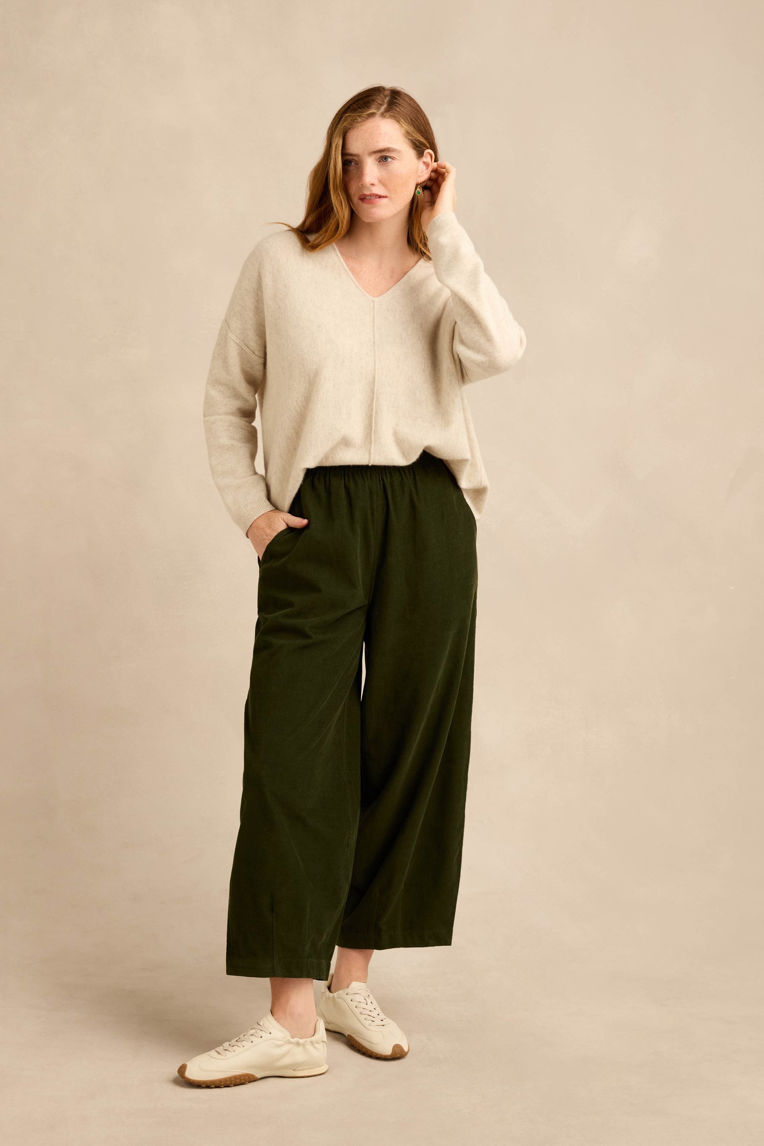 A woman stands against a beige background wearing a beige sweater, the Forest Pinwale Cord Bubble Trousers in a relaxed dark green fit, and white sneakers, with one hand in her pocket and the other touching her hair.