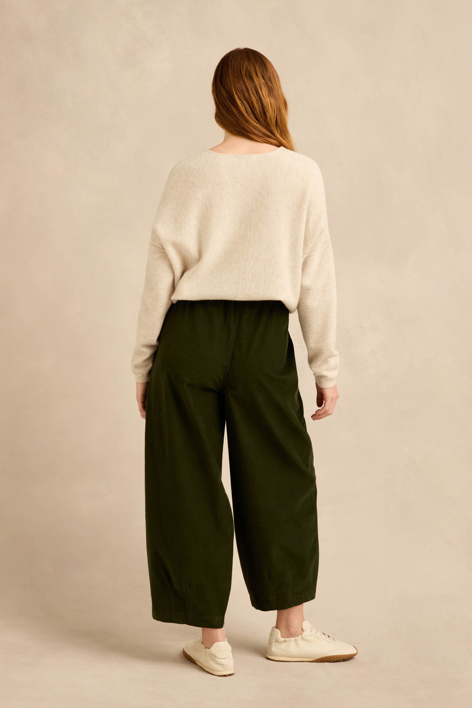 A person stands with their back to the camera, wearing a cream sweater and the Forest Pinwale Cord Bubble Trousers in dark green, featuring a relaxed fit, paired with white sneakers against a plain beige background.
