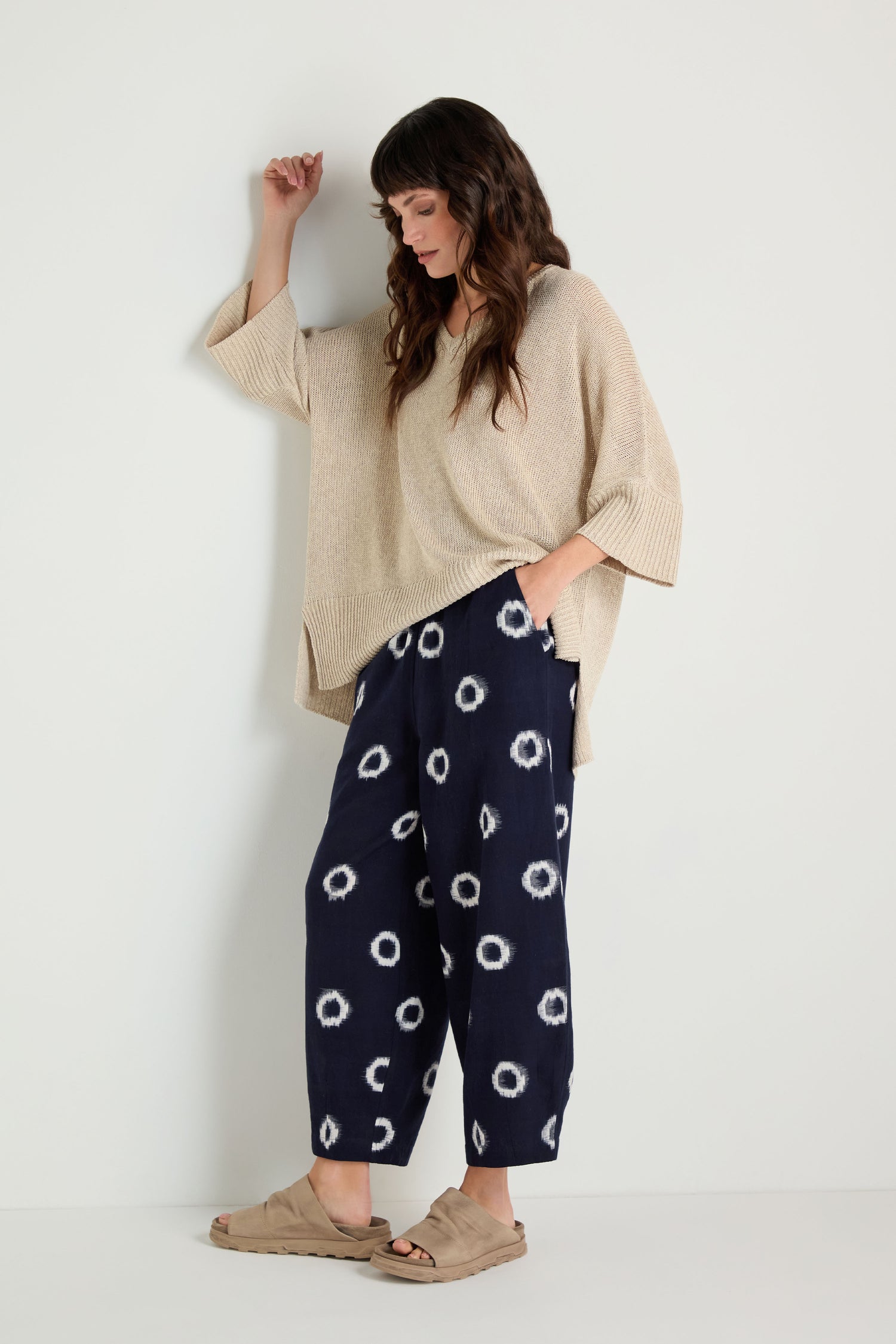 A woman stands against a white wall wearing a beige oversized sweater, Ikat Circles Cotton Bubble Trousers featuring an indigo ecru circular design, and tan slide sandals.