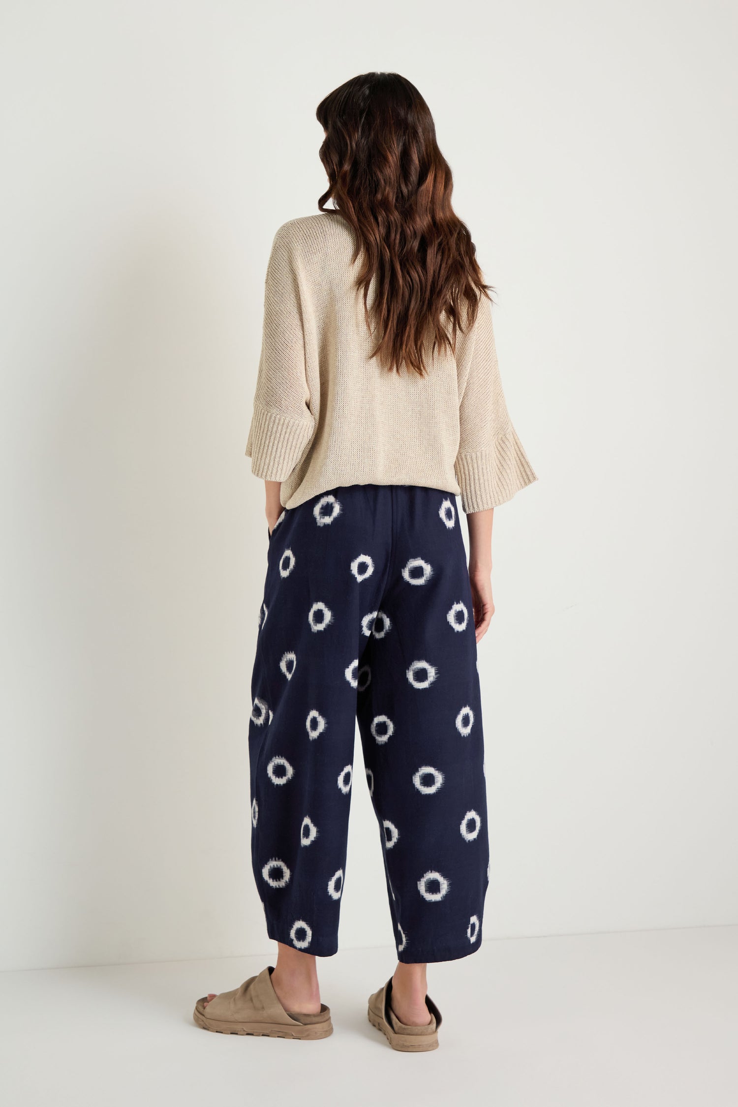 A person stands facing away, wearing beige and indigo Ikat Circles Cotton Bubble Trousers, a beige sweater, and beige sandals against a plain white background.