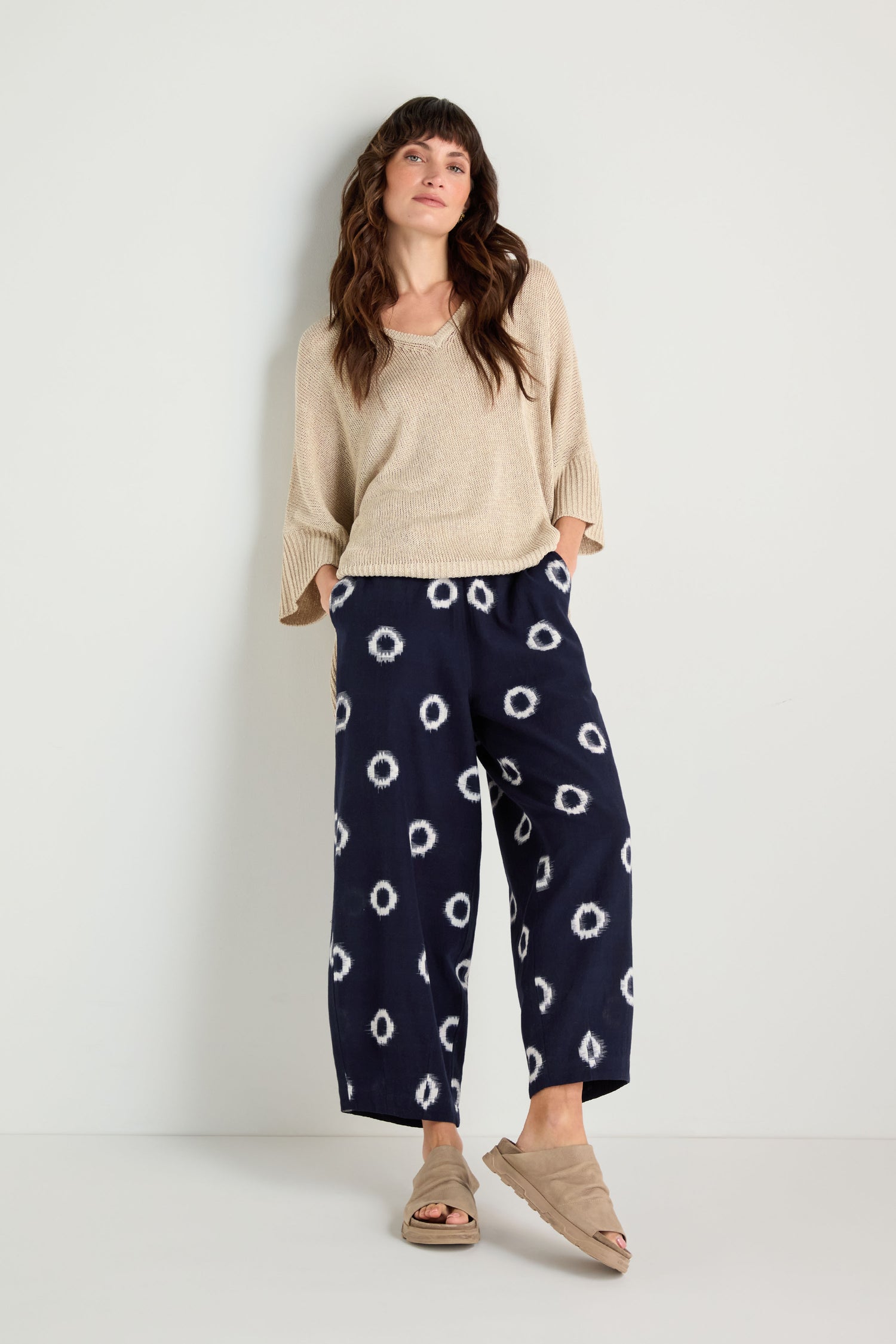 A woman stands against a white wall wearing a beige sweater, Ikat Circles Cotton Bubble Trousers, and beige sandals.