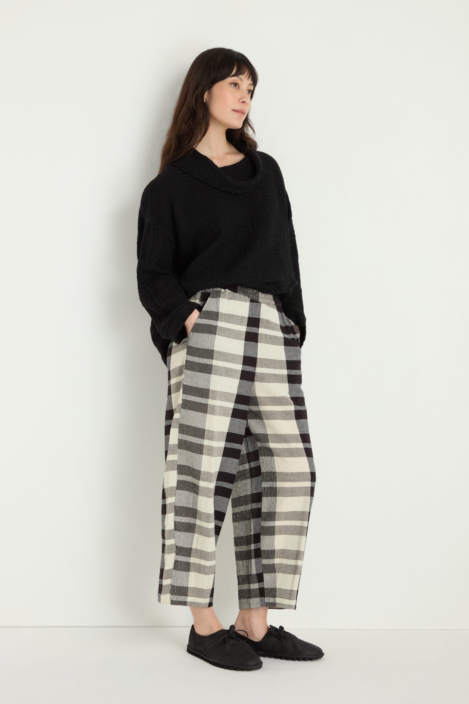 A woman stands against a white wall wearing a black sweater, Monochrome Crinkle Check Bubble Trouser with an elasticated waist, and black shoes.
