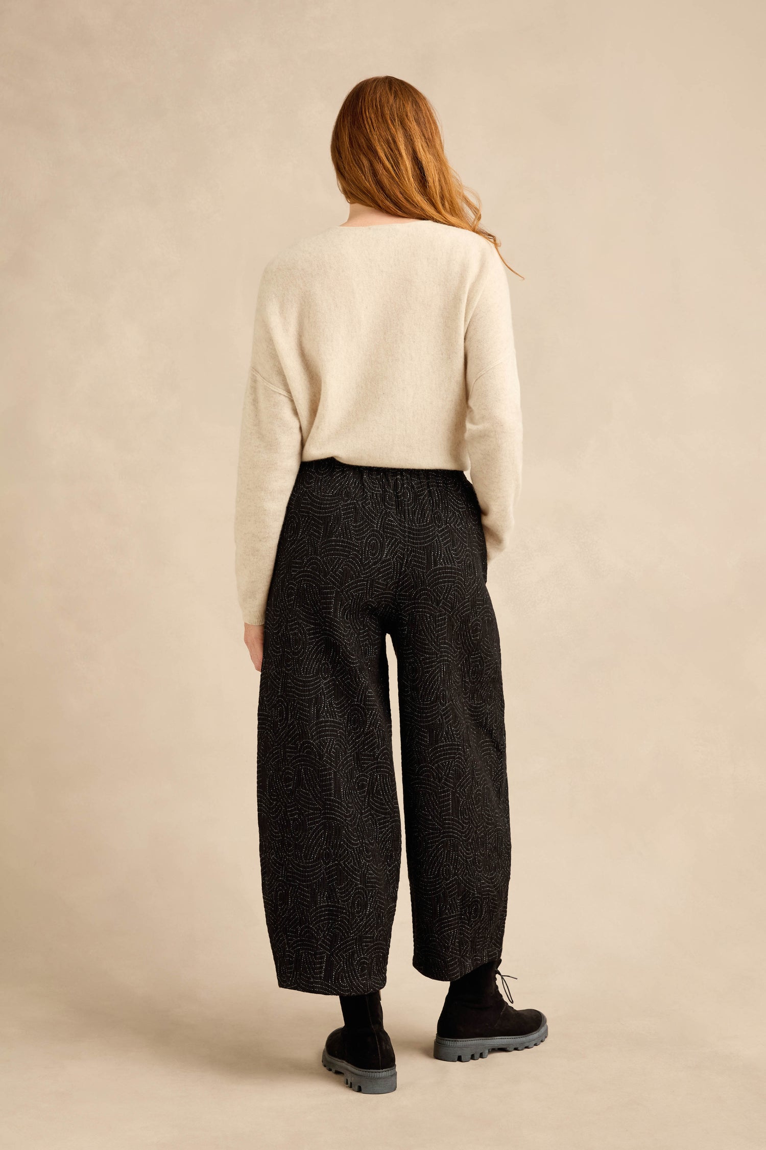 A person with long hair, wearing a cream sweater, the Stitched Jacquard Bubble Trouser in black, black socks, and black shoes, stands facing away against a beige background.
