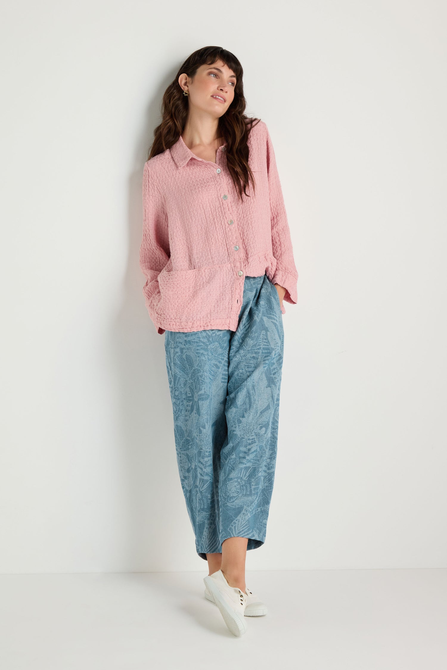 A woman stands against a white wall wearing a pink textured button-up shirt, the Floral Jacquard Cotton Bubble Trouser in blue with a cropped bubble shape, and white sneakers.