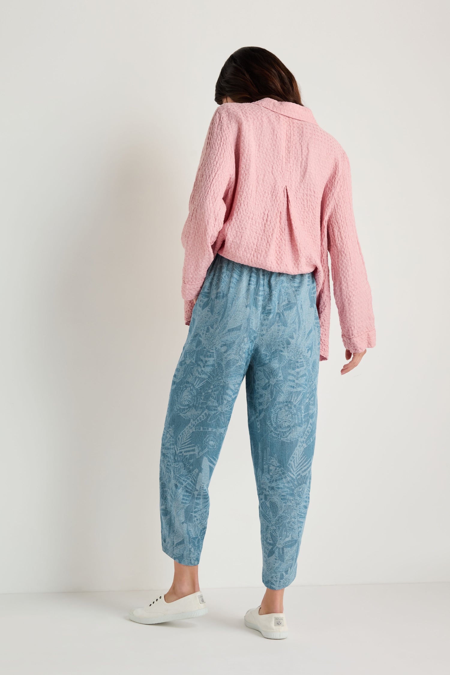 A person stands facing away, wearing a textured pink shirt, Floral Jacquard Cotton Bubble Trouser, and white lace-up shoes against a plain white background.