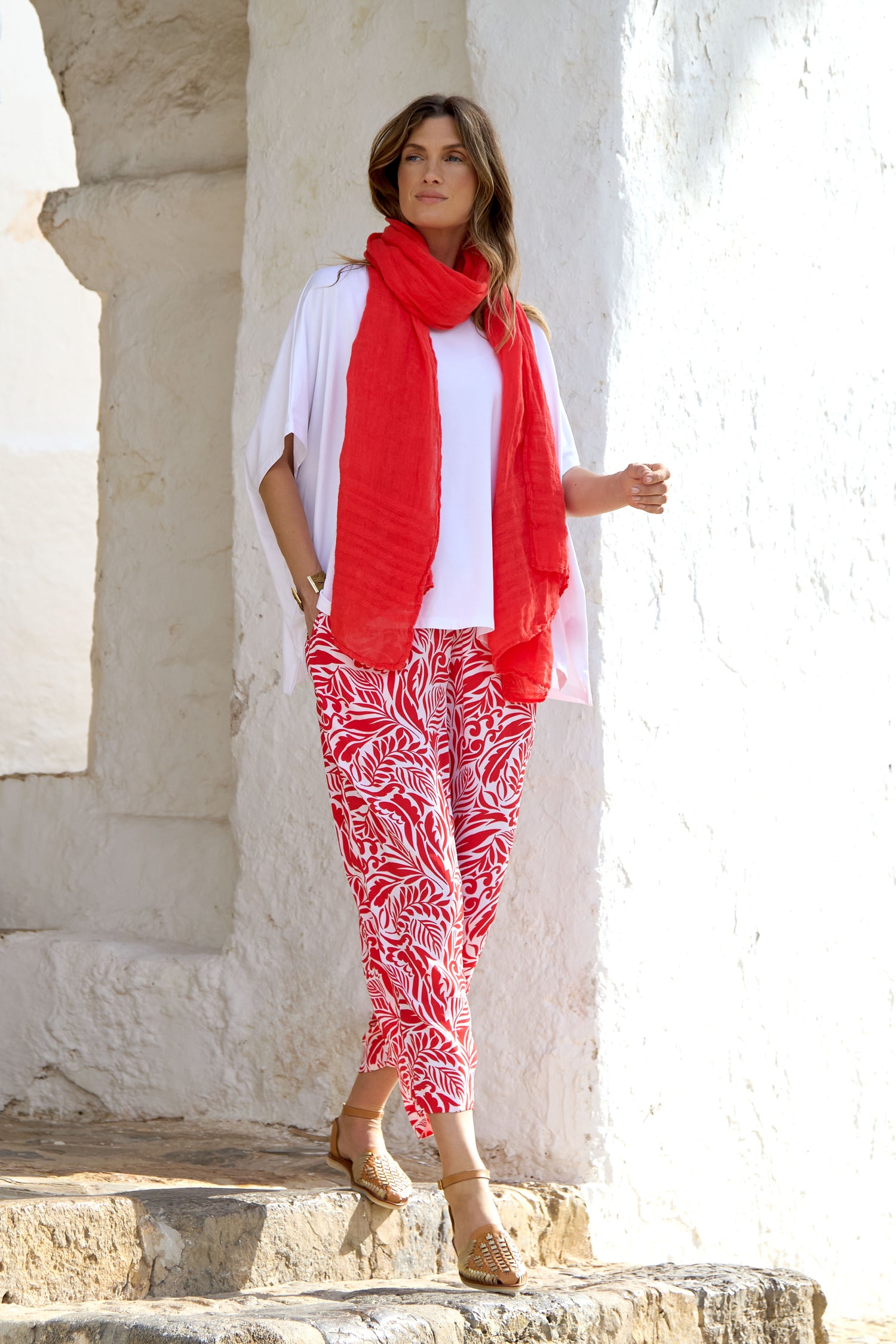A woman embracing a summer vibe poses on stone steps against a light wall, dressed in a white top, red scarf, vibrant Tropical Fusion Bubble Trouser, and brown sandals.