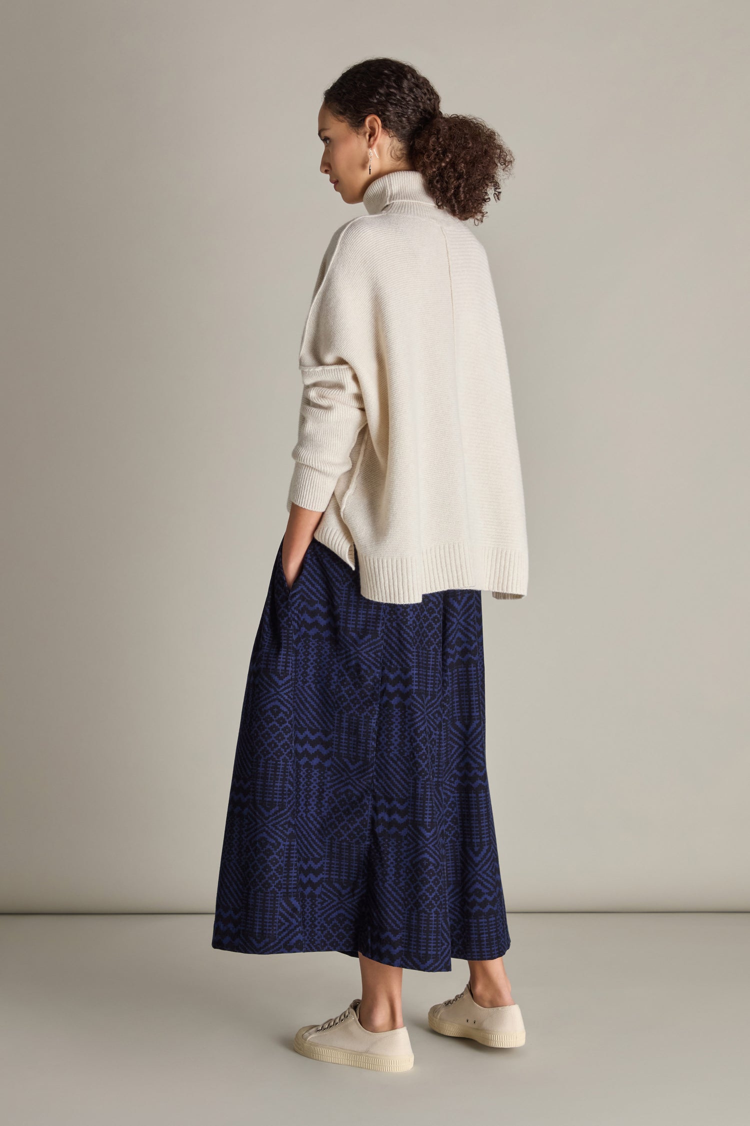 A person with curly hair is standing sideways, wearing a cream turtleneck sweater, Mono Ikat Printed Cord Trouser that gives a tailored look, and white sneakers, against a plain background.