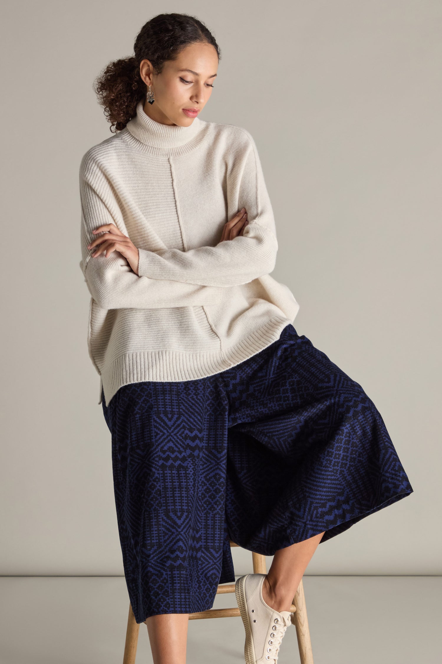 A woman with curly hair is wearing a beige turtleneck sweater and Mono Ikat Printed Cord Trousers. She sits on a stool with her arms crossed and a neutral expression, set against a plain background, showcasing an elegant ensemble.