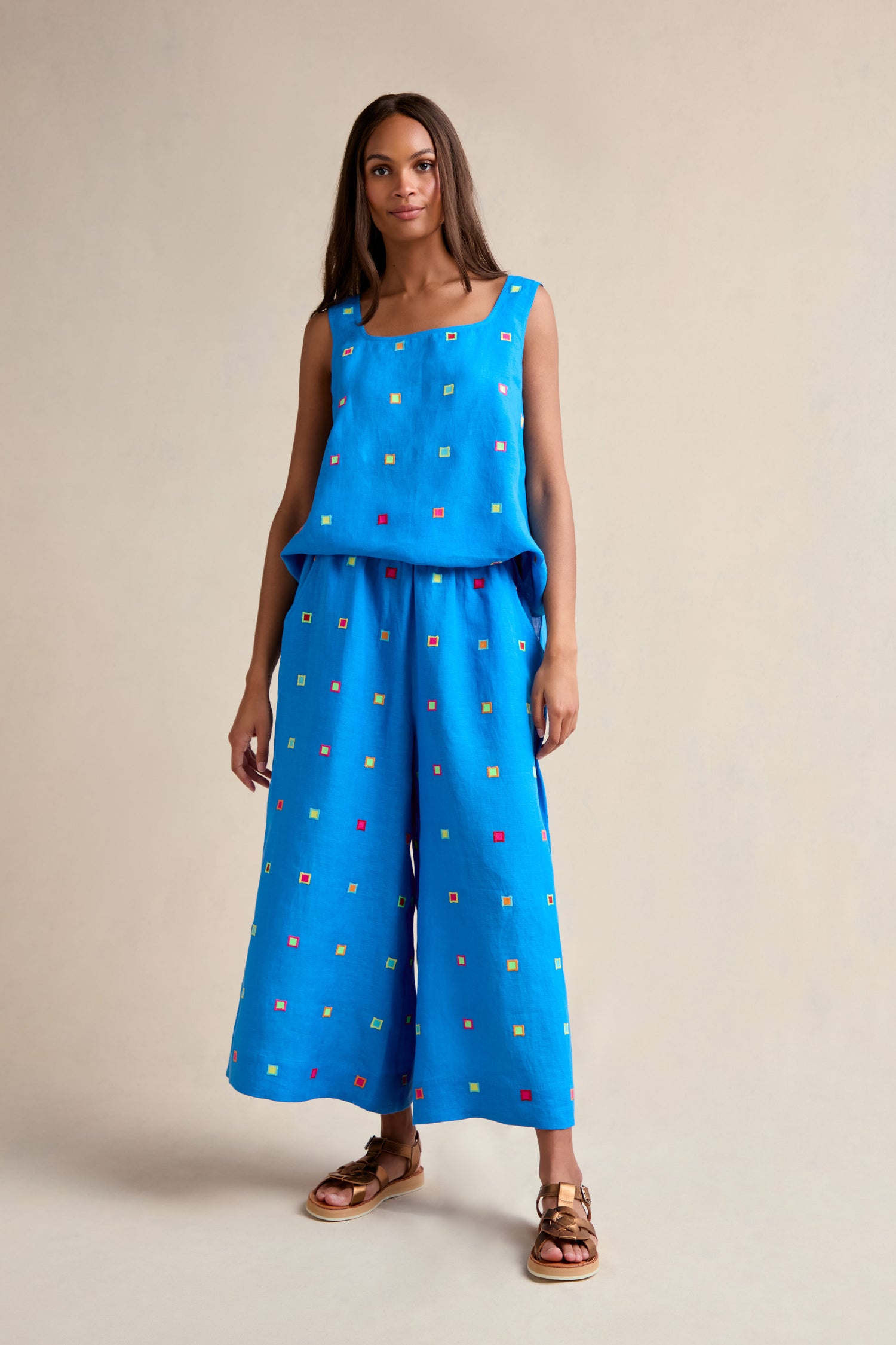 A woman stands against a beige background wearing Embroidered Squares Linen Wide Trousers—a loose, cropped linen style with small embroidered squares—paired with a sleeveless blue top.