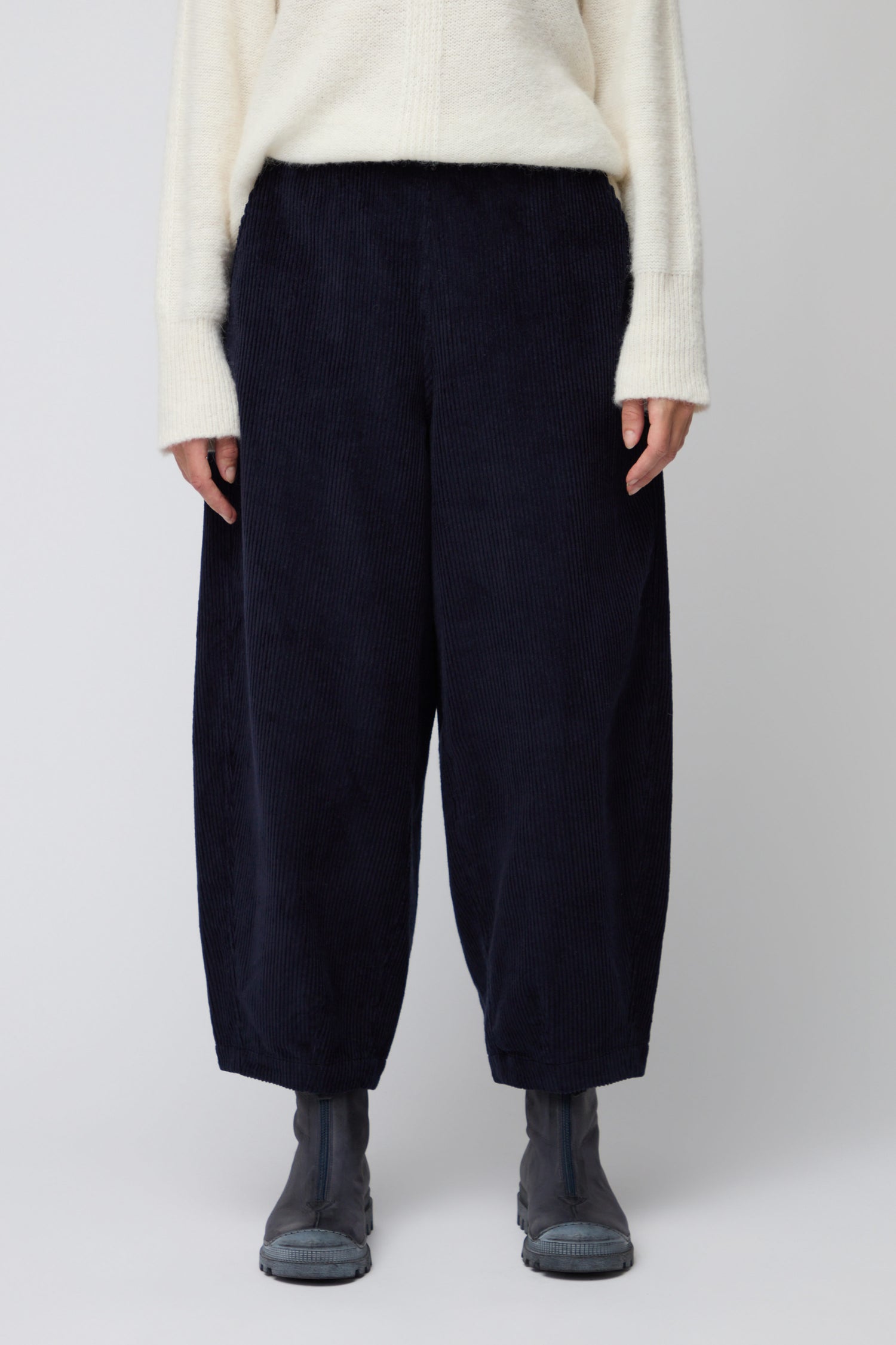Person wearing a cream sweater, Jumbo Cord Bubble Trousers in navy, and gray ankle boots stands against a plain background.
