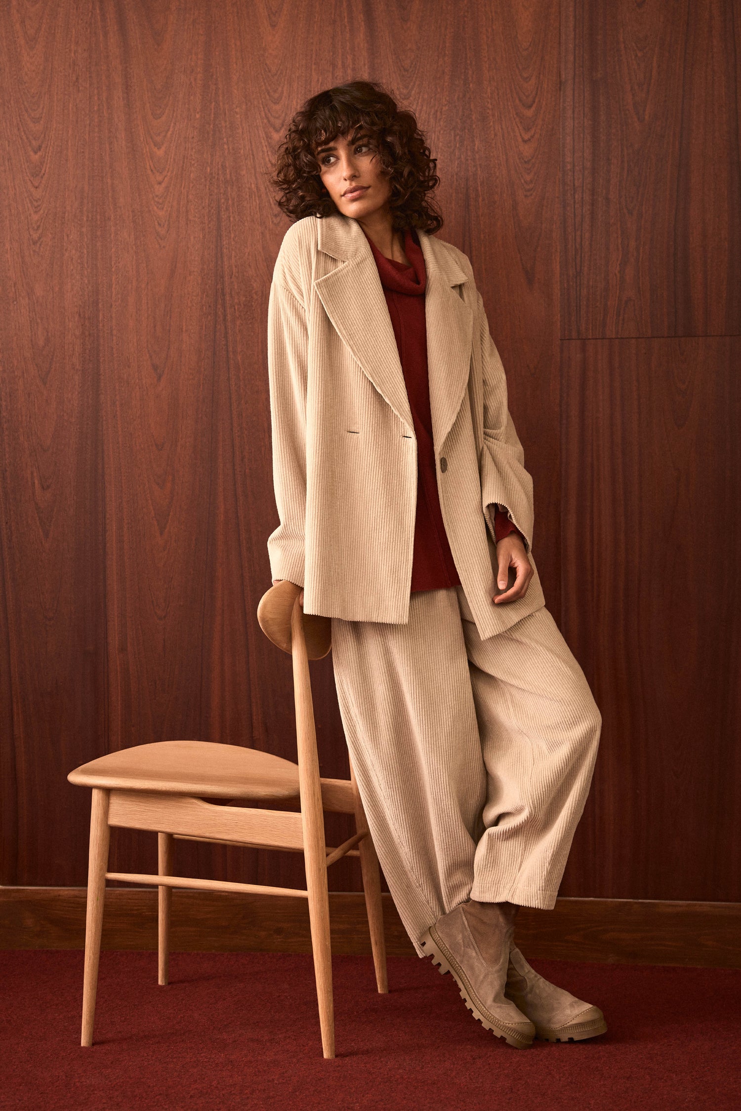 Person with curly hair leans against a wooden chair, wearing a beige suit featuring the Ecru Jumbo Cord Bubble Trousers, a red top, and beige boots, in a room with wood-paneled walls and a red carpet.