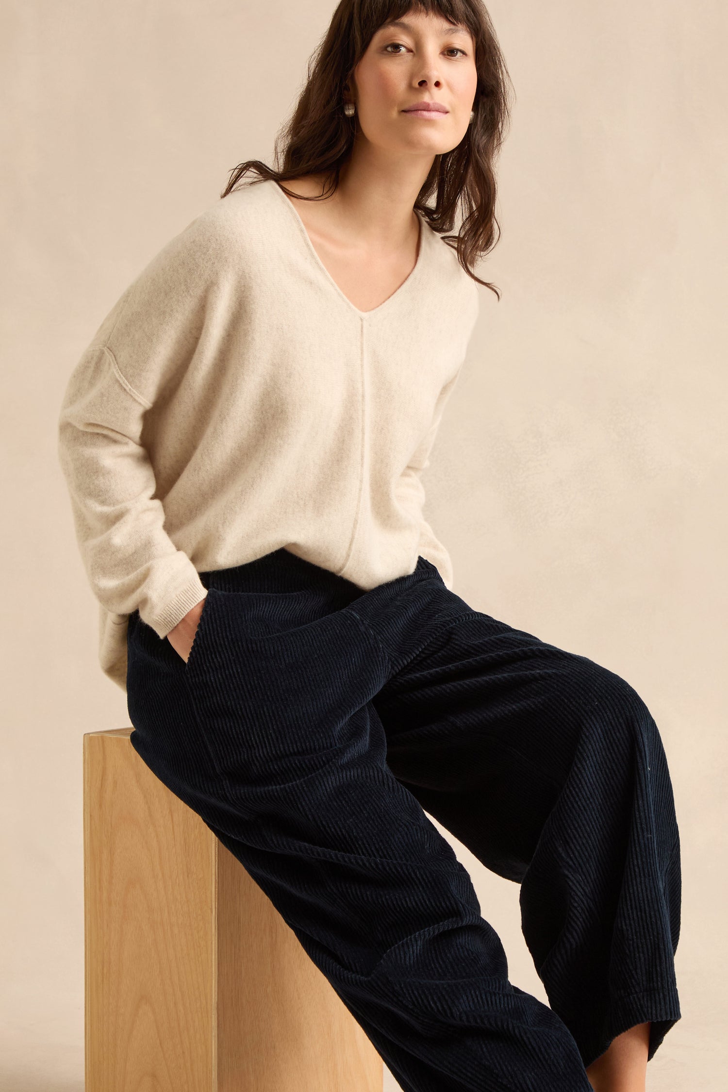 A woman in a cream pure cotton V-neck sweater and navy jumbo cord bubble trousers sits on a wooden block against a plain beige background.
