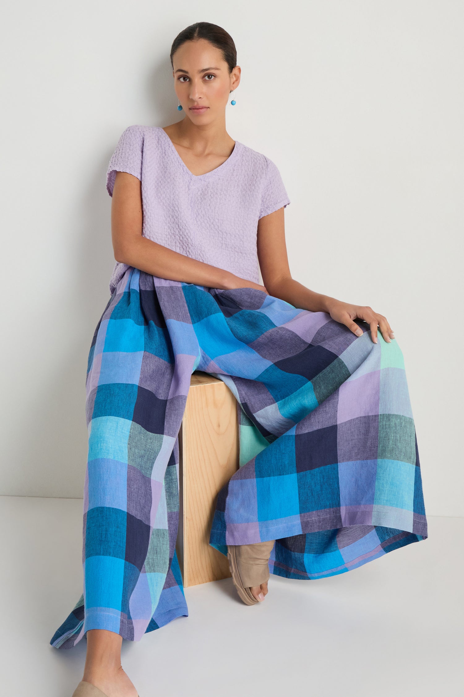 A woman sits on a wooden block, wearing a light purple textured top and blue and purple Cross Dye Check Linen Wide Trousers, against a plain white background.