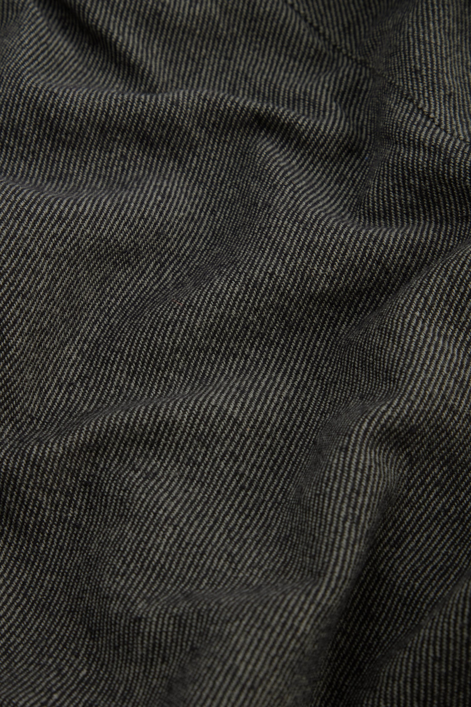 Close-up of dark gray and black pinstripe fabric with subtle folds and texture, highlighting the refined finish characteristic of the Overdyed Twill Barrel Trouser.
