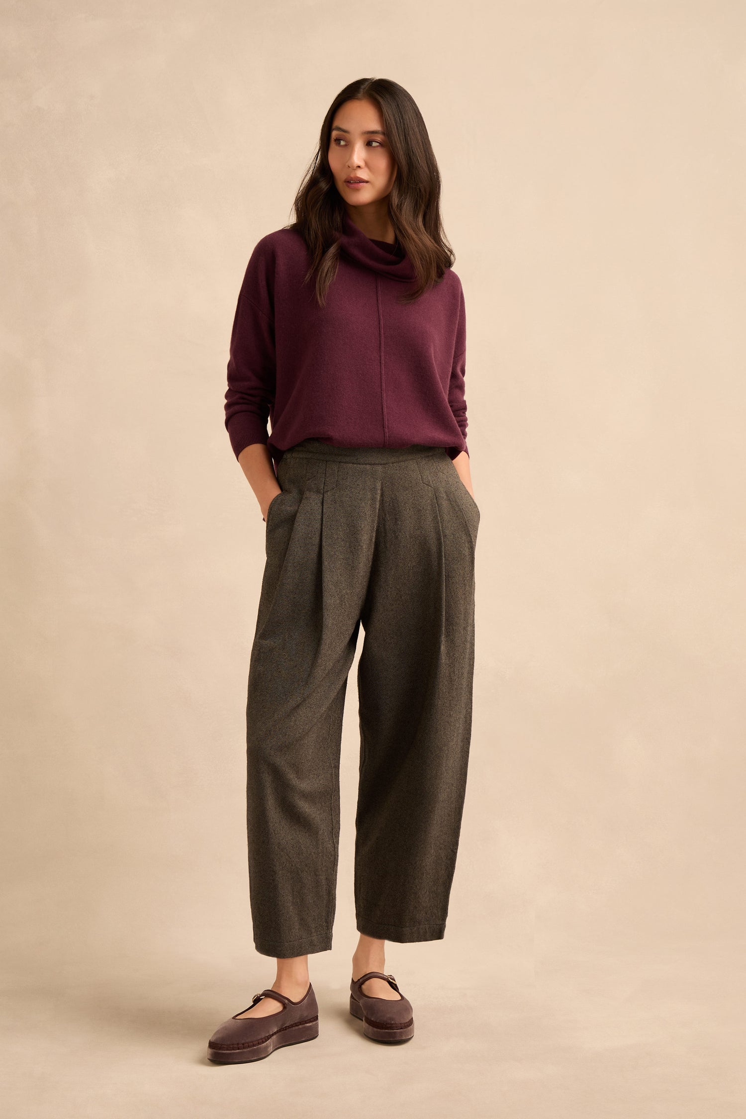 A woman stands against a neutral background, wearing a burgundy sweater and the Overdyed Twill Barrel Trouser in loose gray pleats, paired with mauve shoes. She has her hands in her pockets and looks slightly to the side.