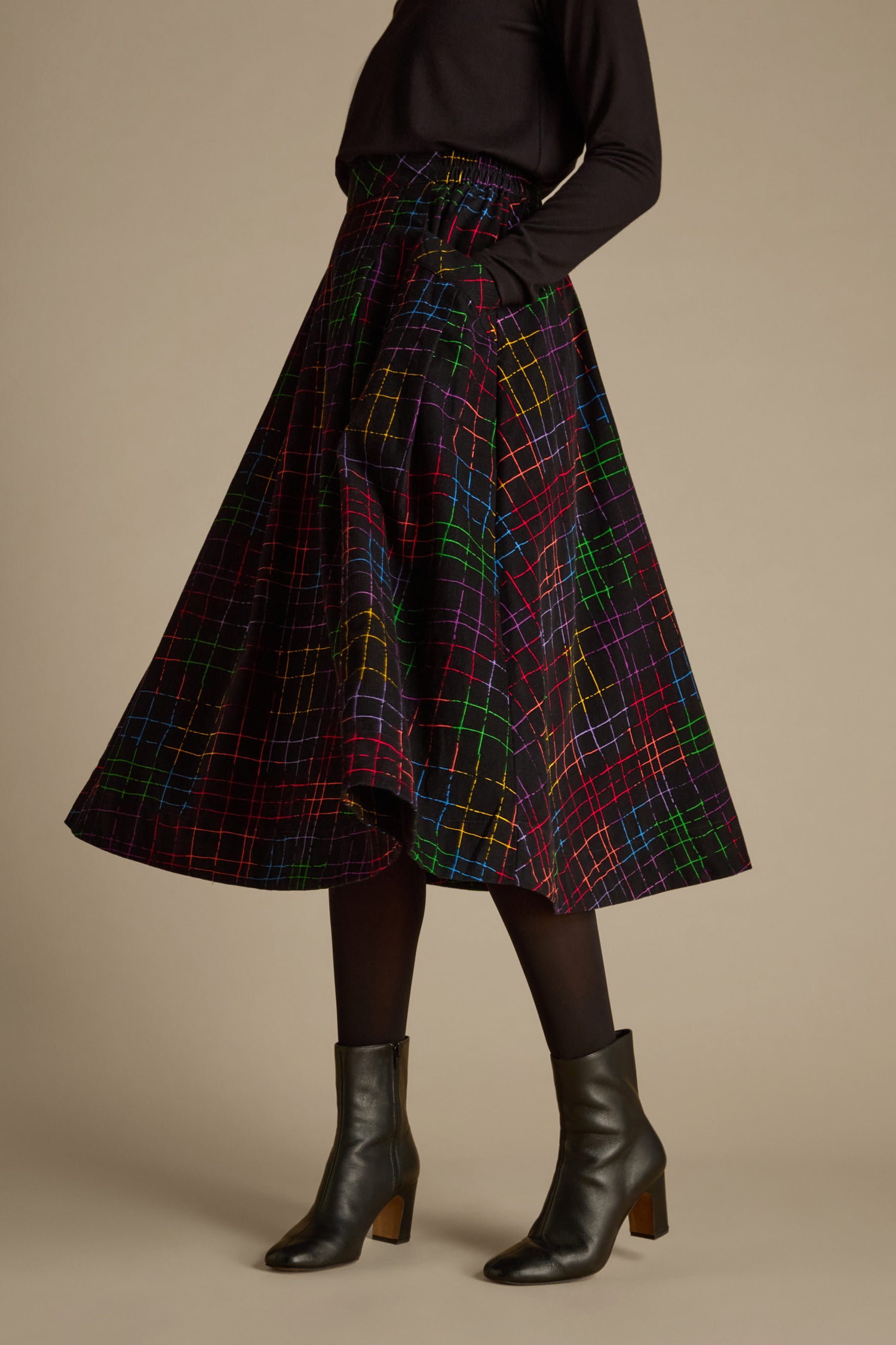 A person wearing a black top and black ankle boots stands against a plain background, showcasing the Stitched Check Cord Skirt with its vibrant check print and paired with black tights.