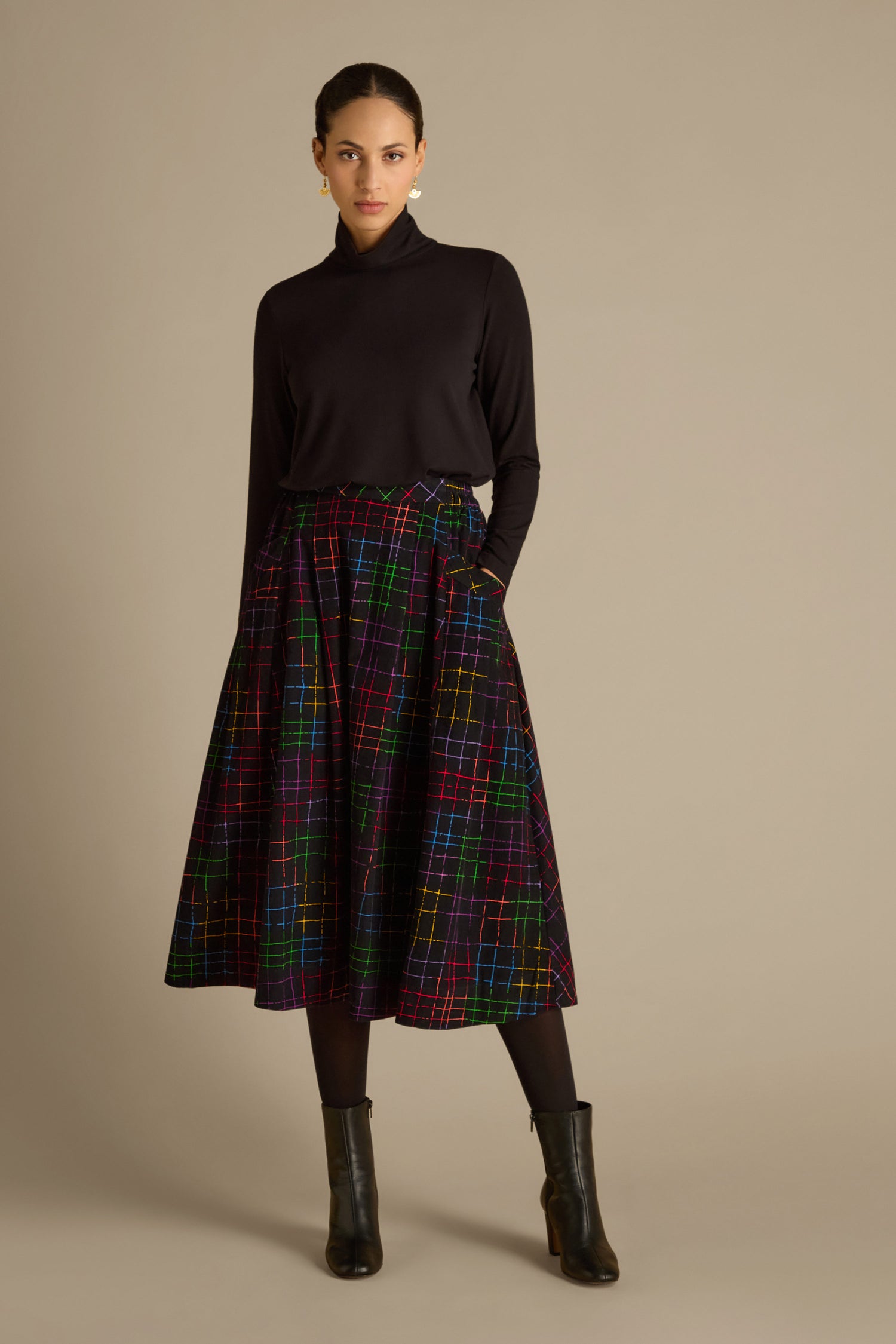 A person stands confidently, dressed in a black turtleneck paired with the Stitched Check Cord Skirt. Complementing the ensemble, they wear black tights and sleek ankle boots against a plain background.