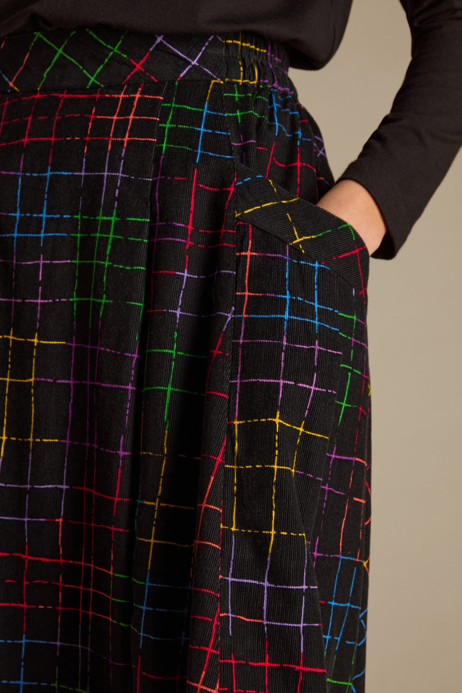 A person wearing a black top and the Stitched Check Cord Skirt, which features a vibrant grid pattern, casually places a hand in the pocket.