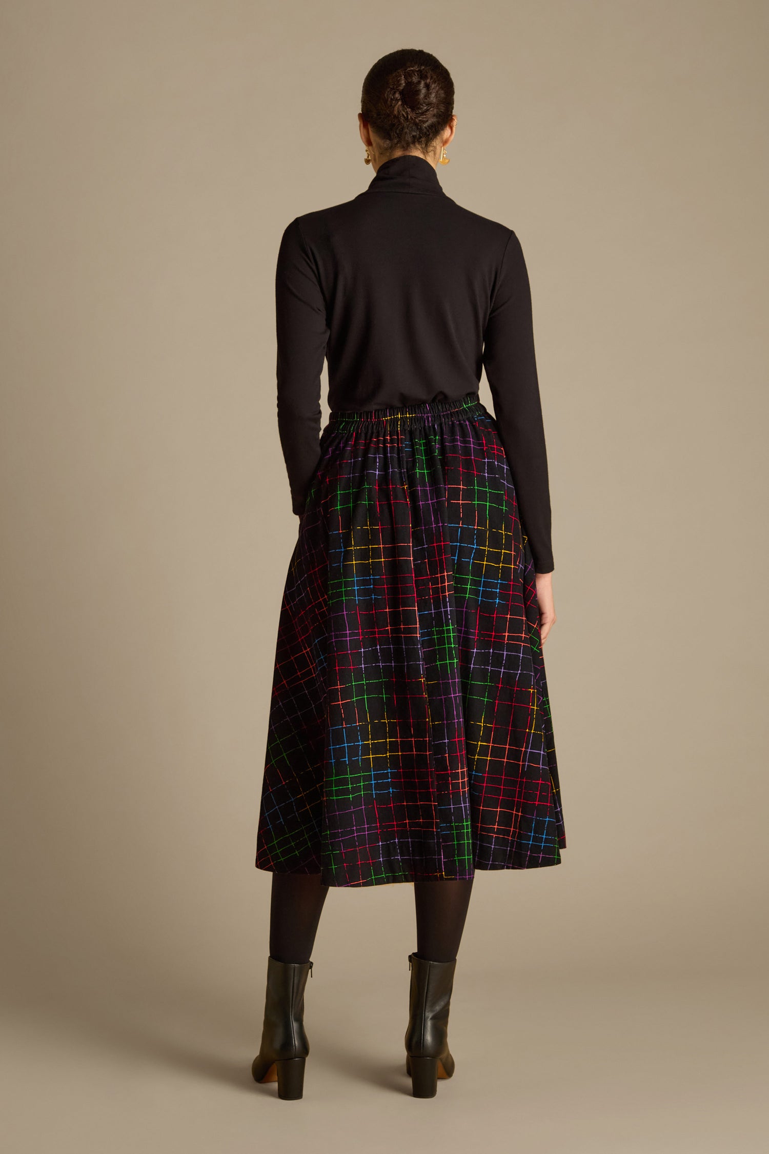 A person with braided hair stands facing away, wearing a black turtleneck, the Stitched Check Cord Skirt, black tights, and black ankle boots against a neutral background.