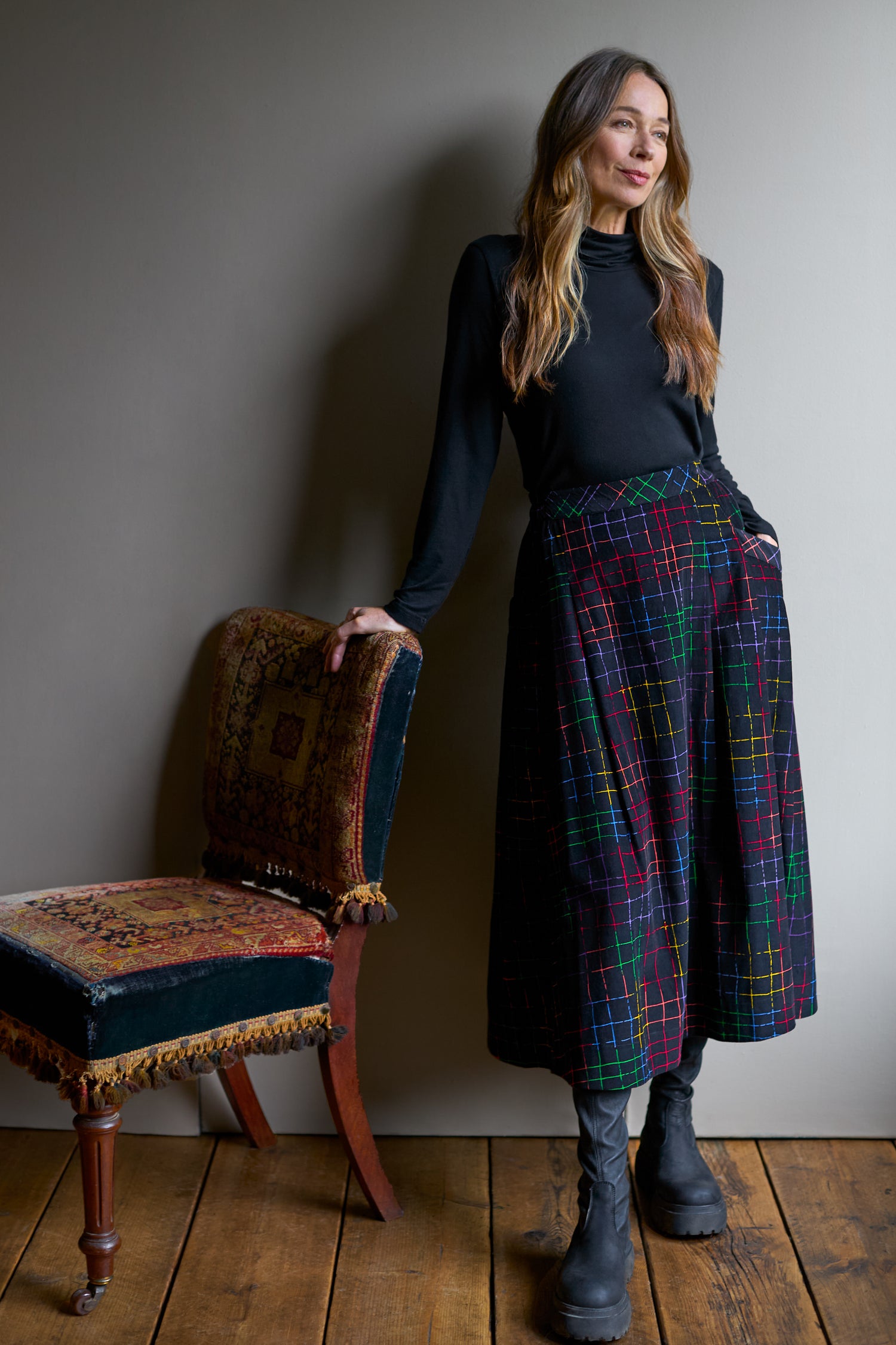 A person is beside a chair, dressed in a black top and black boots. Their Stitched Check Cord Skirt, with its vibrant hues and A-line silhouette, brings flair to the simple wooden floor and plain wall backdrop.