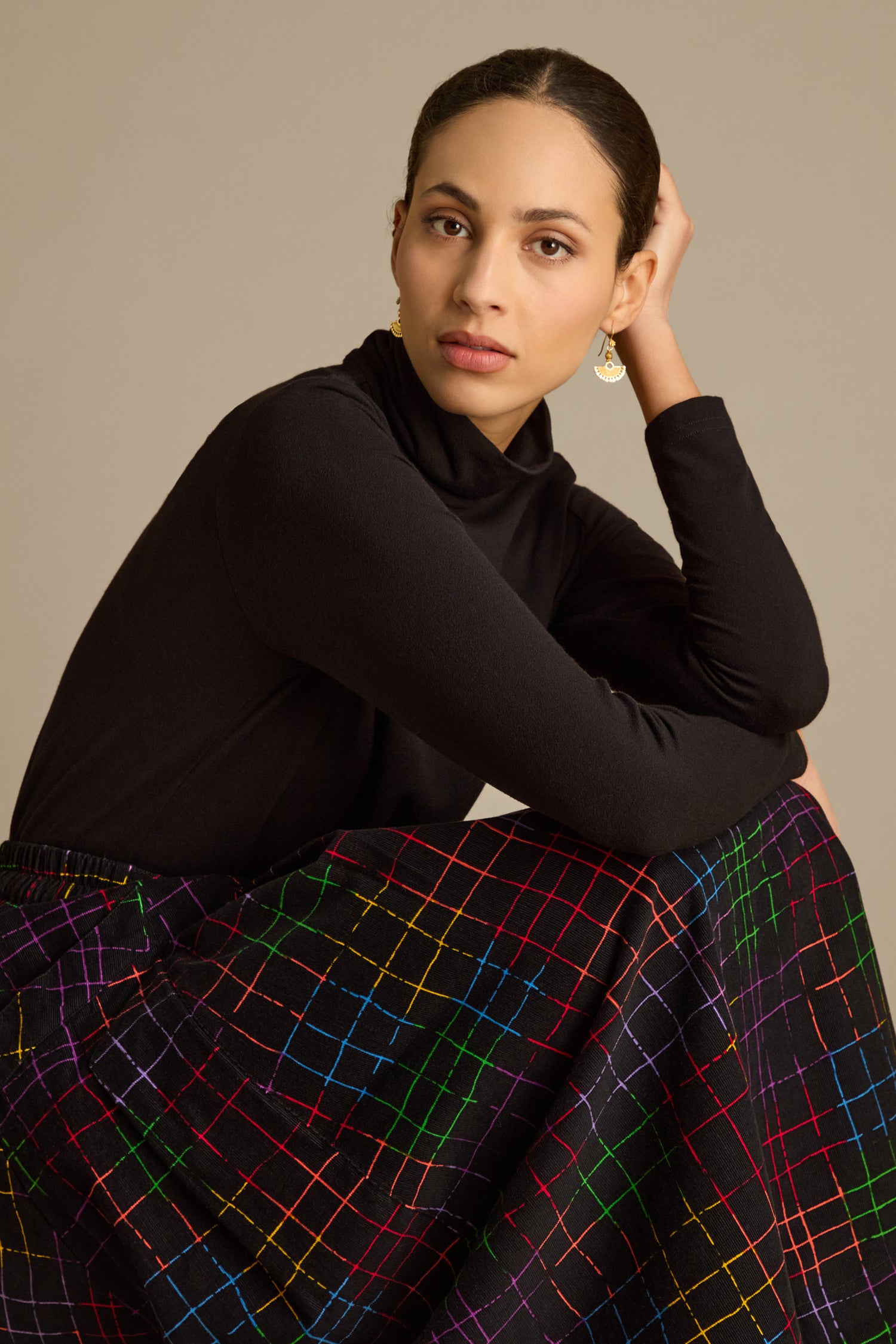 A person wearing a black turtleneck and the vibrant Stitched Check Cord Skirt sits with one arm resting on their knee, against a neutral background.
