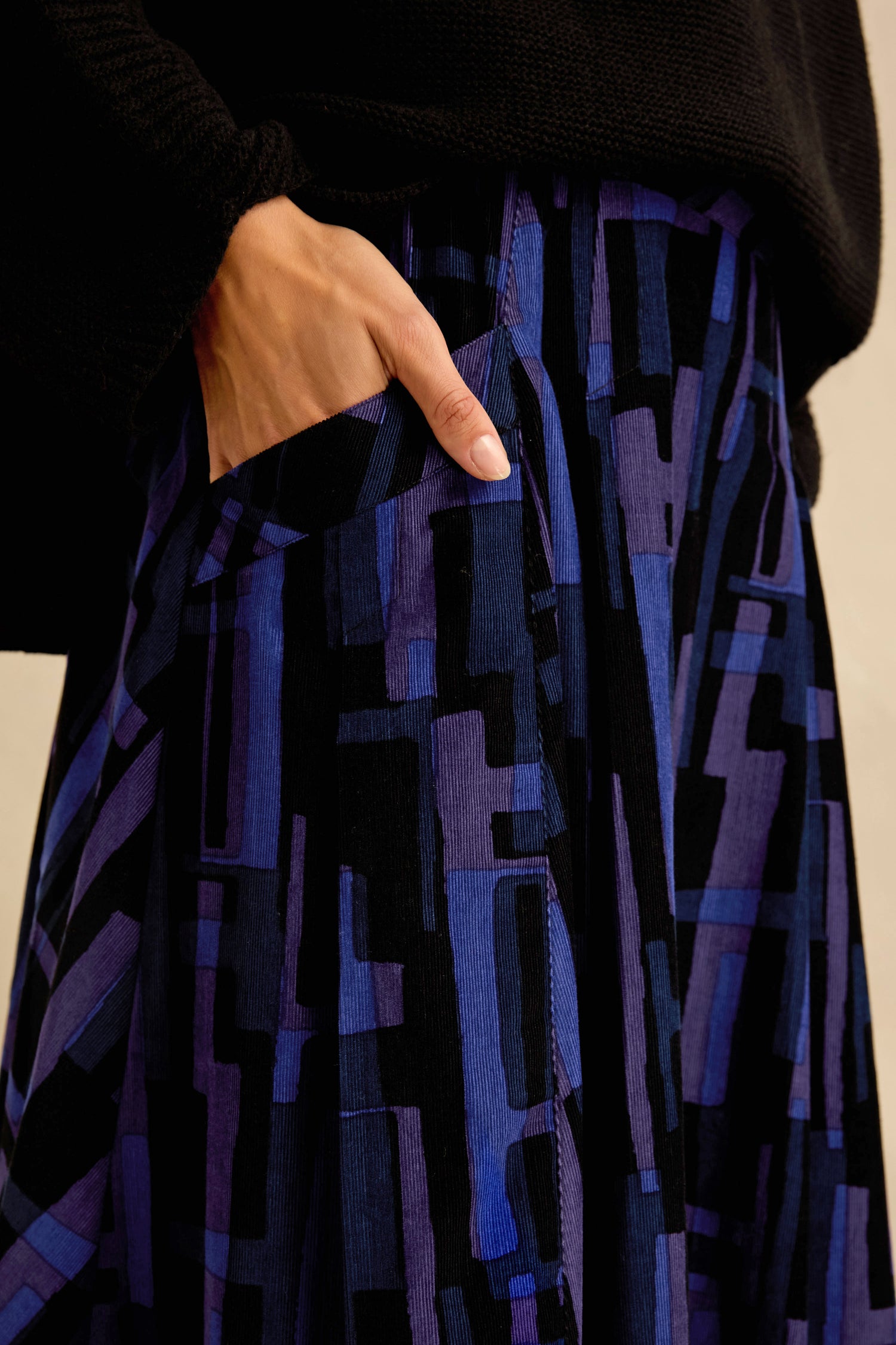 A person wearing a black sweater and the Layered Geometric Cord Skirt, featuring a blue and black geometric pattern in an A-line corduroy style, with one hand in the skirt pocket.