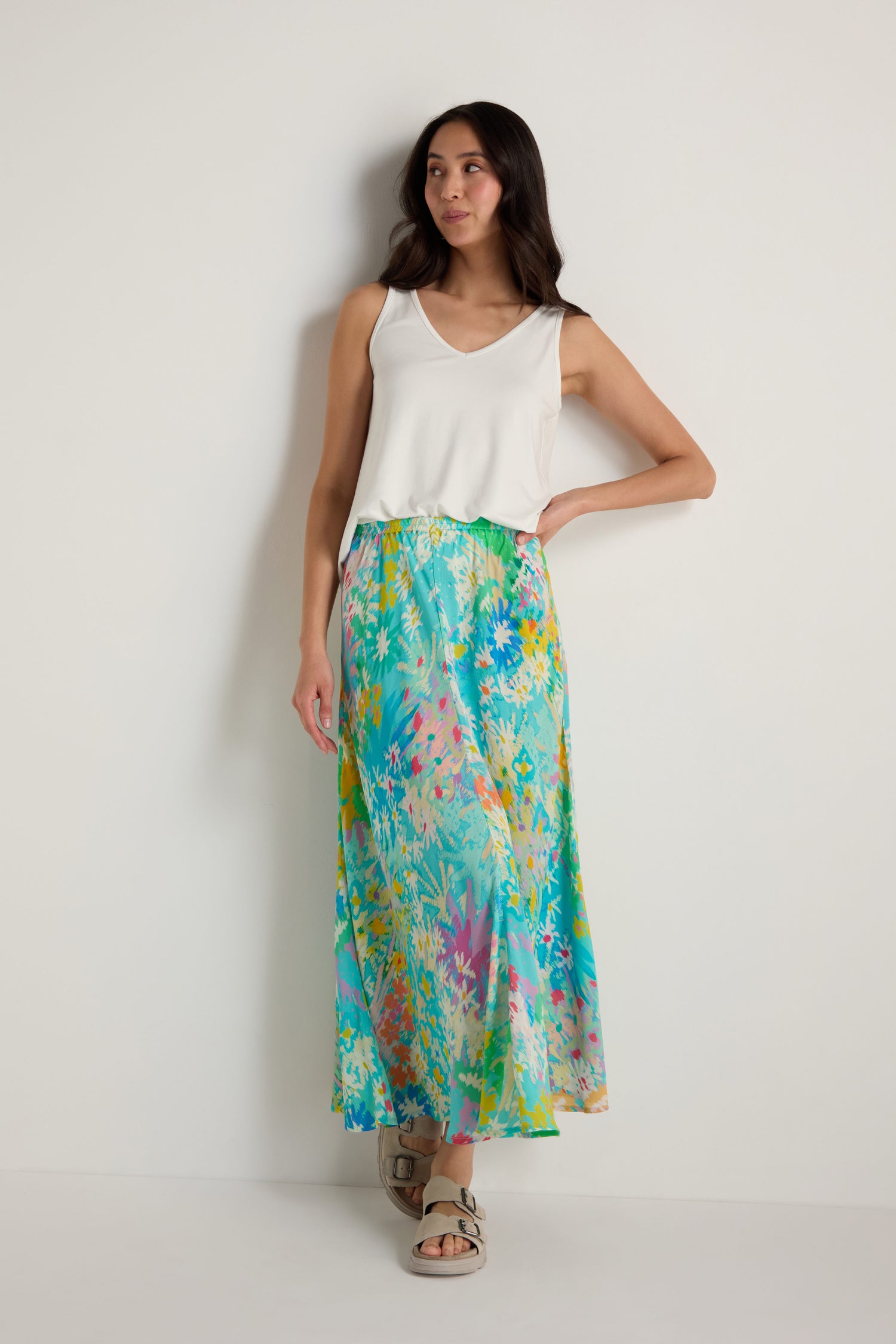 A woman stands against a plain white wall wearing a white sleeveless top, beige sandals, and the Floral Oasis Bias Skirt with its colorful floral pattern.