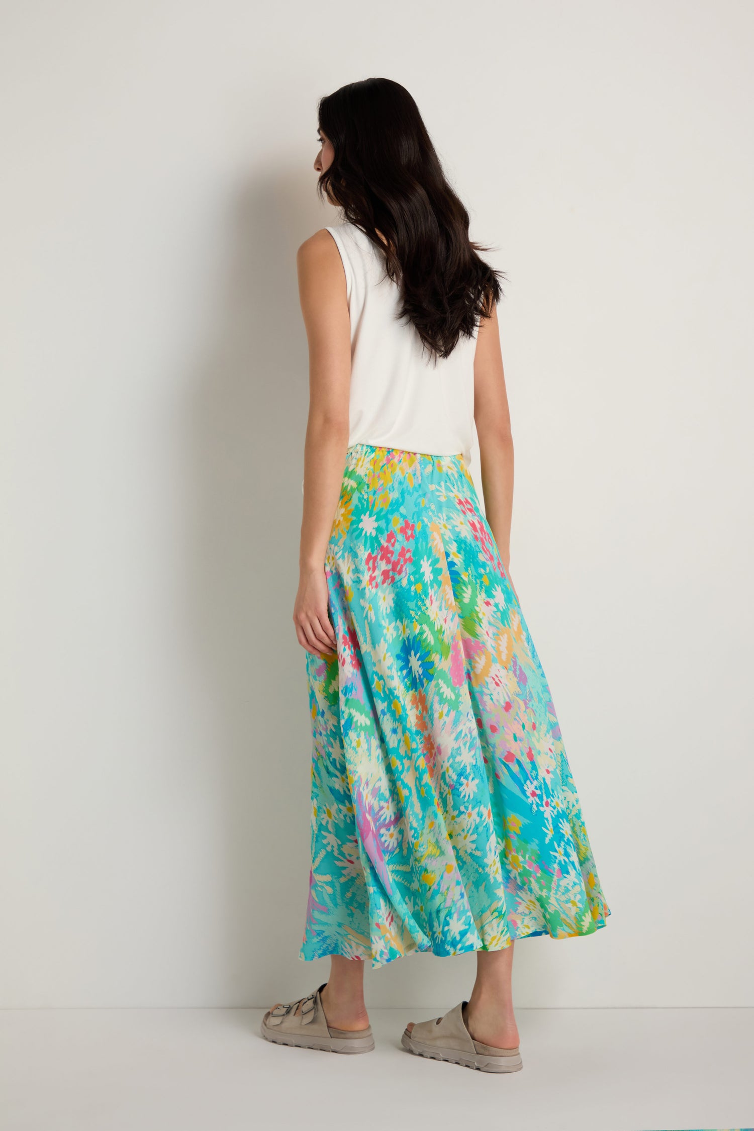 A woman stands facing away, wearing a white sleeveless top and the Floral Oasis Bias Skirt—a colorful floral midi—and beige slip-on shoes.