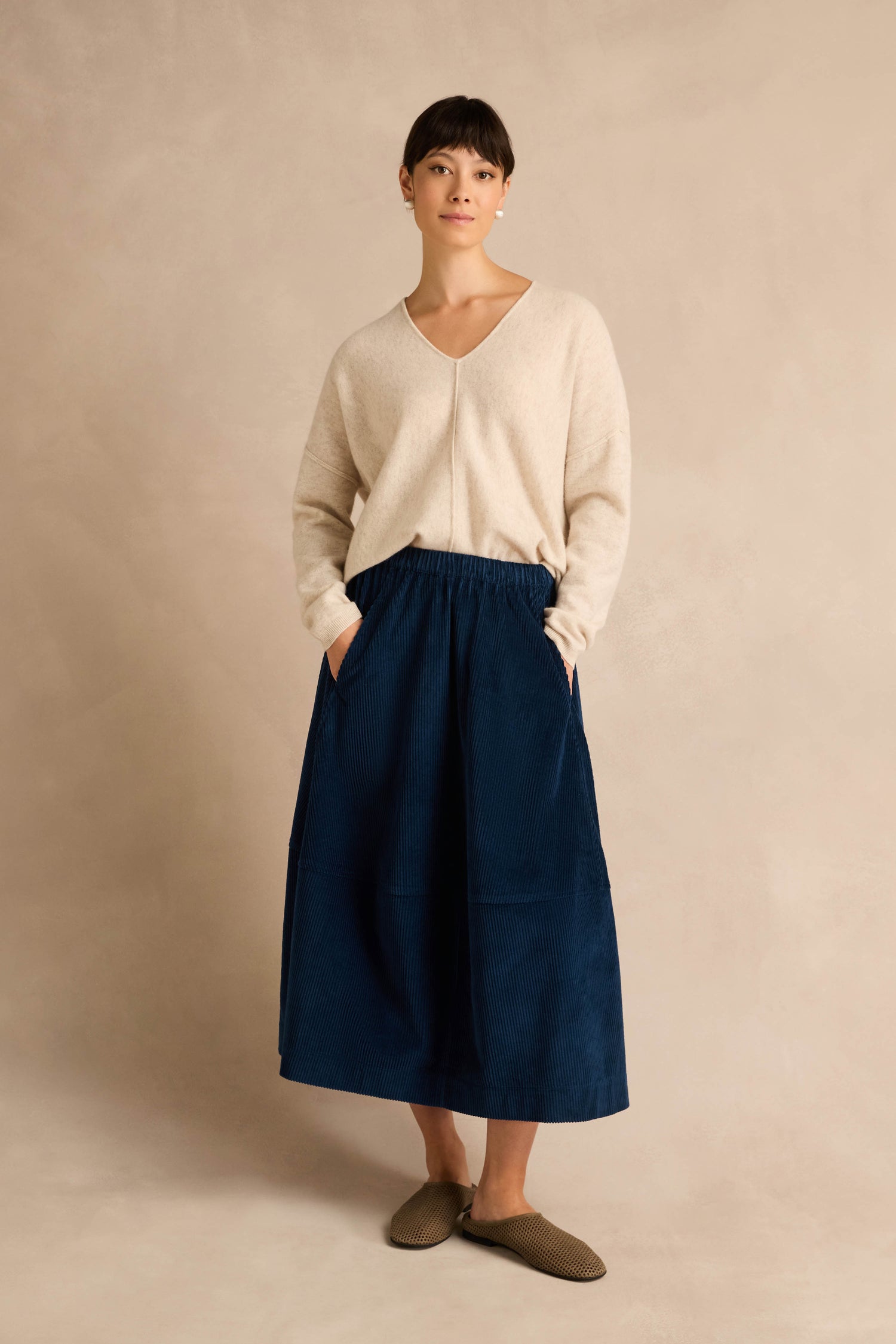 A woman stands against a plain beige background wearing a cream sweater, brown slippers, and the Jumbo Cord Bubble Skirt in navy blue cotton with pockets.