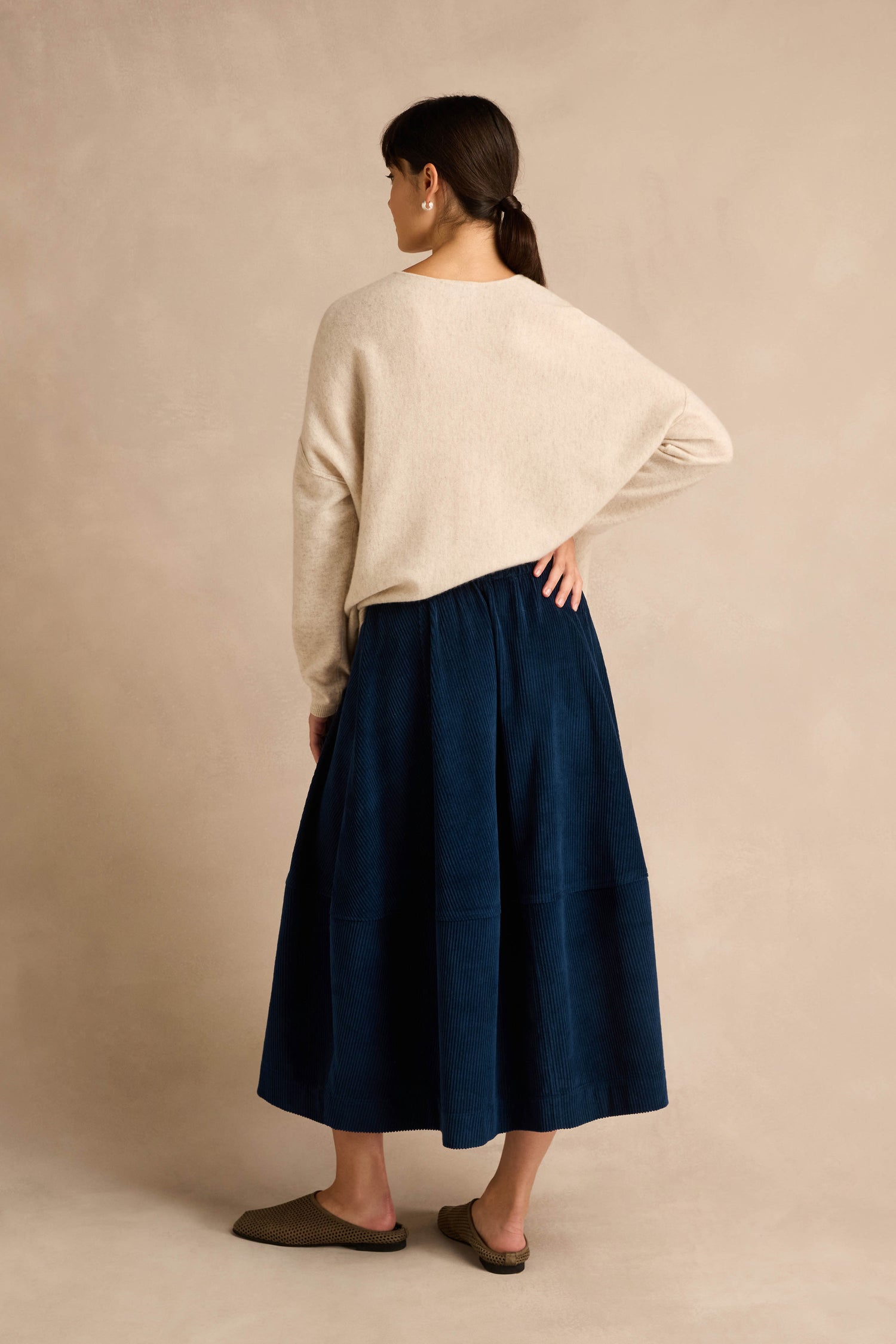 A woman stands facing away, wearing a beige long-sleeve sweater, the Jumbo Cord Bubble Skirt in navy blue with its subtle bubble silhouette and cotton texture, paired with brown slippers, against a plain beige background.