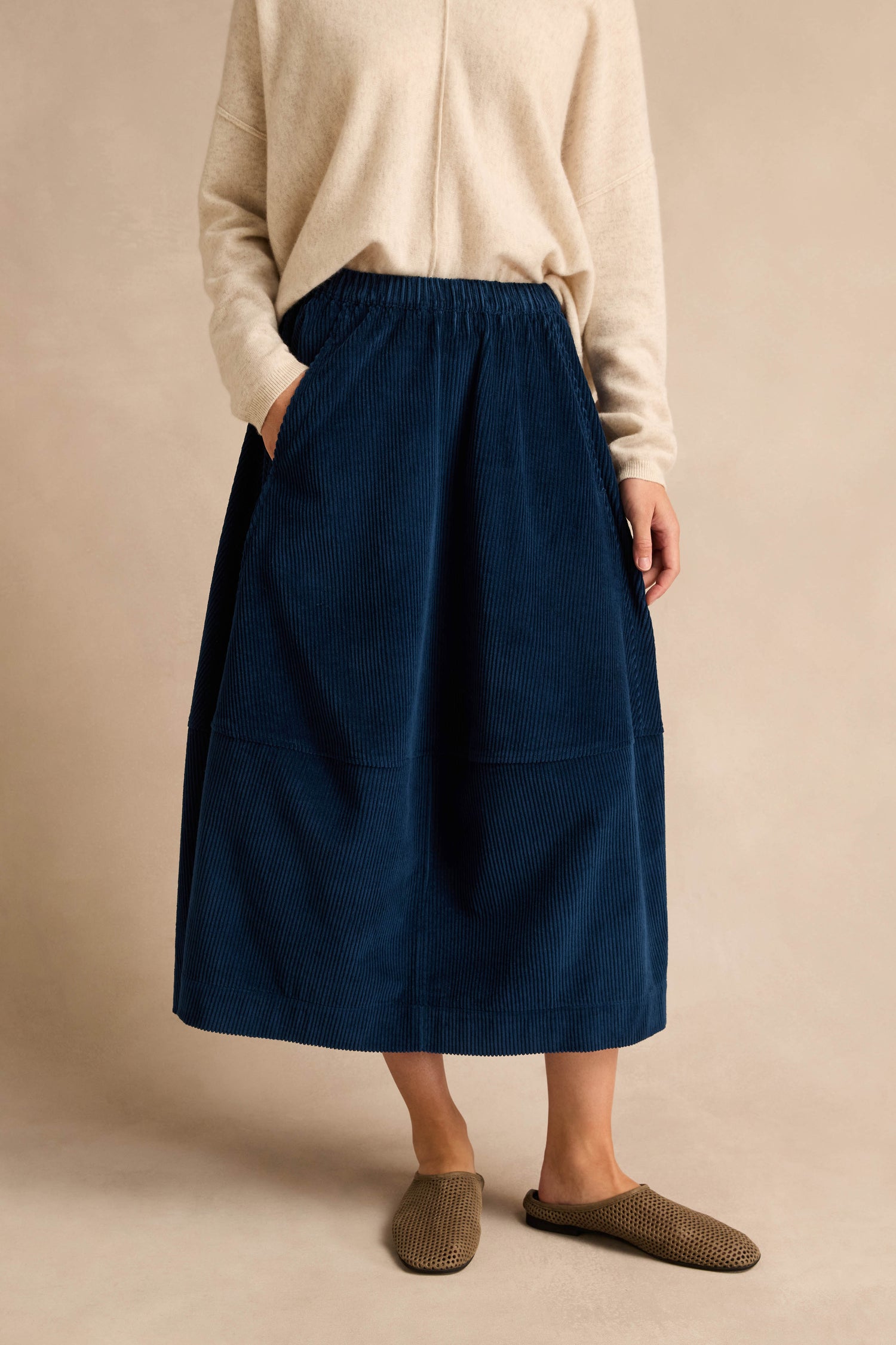 Person wearing a cream long-sleeve top, the Jumbo Cord Bubble Skirt in blue with pockets, and brown slip-on shoes, standing against a neutral background.