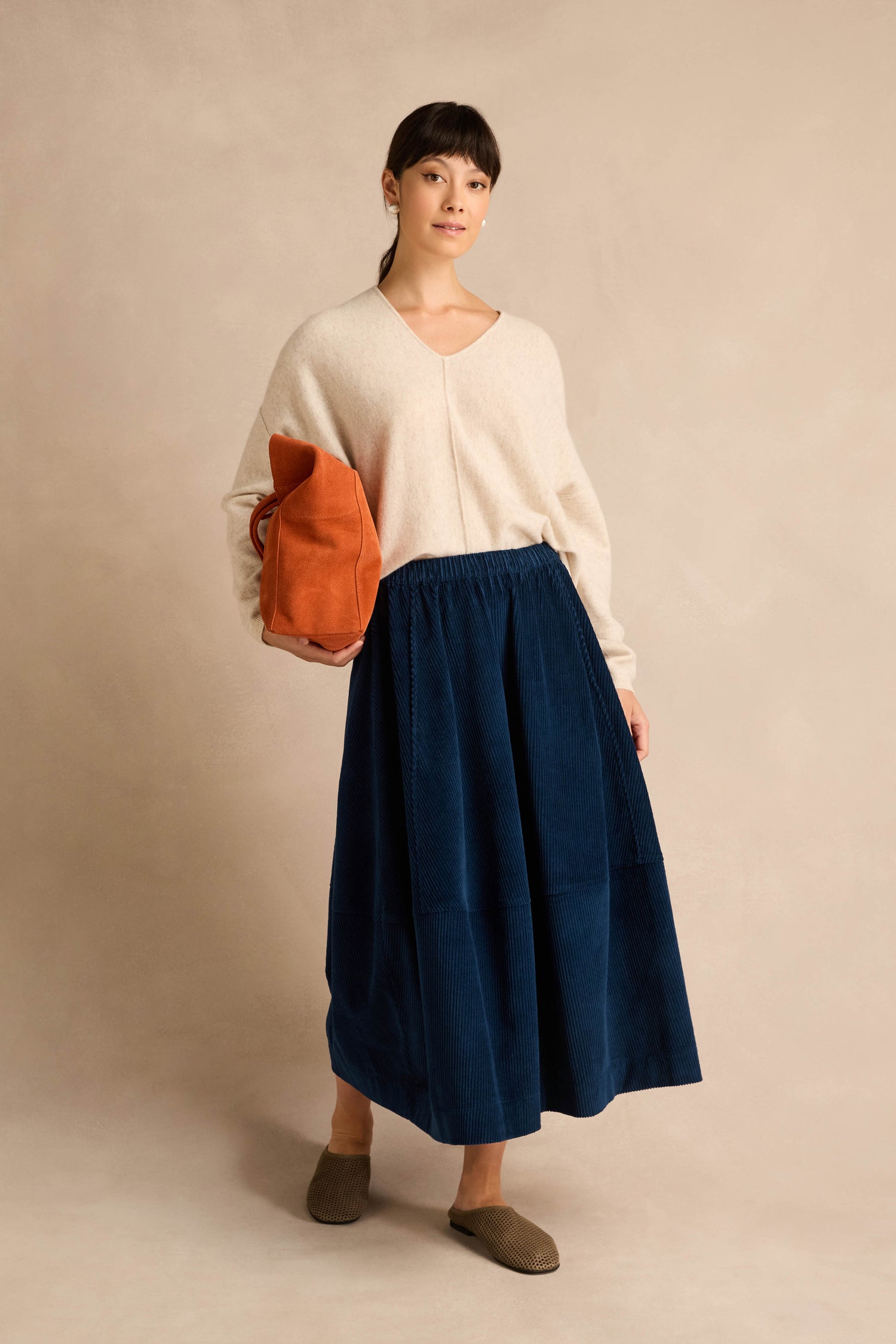A woman stands against a plain backdrop wearing a beige sweater, the Jumbo Cord Bubble Skirt in dark blue cotton, brown shoes, and holding an orange bag under her arm.