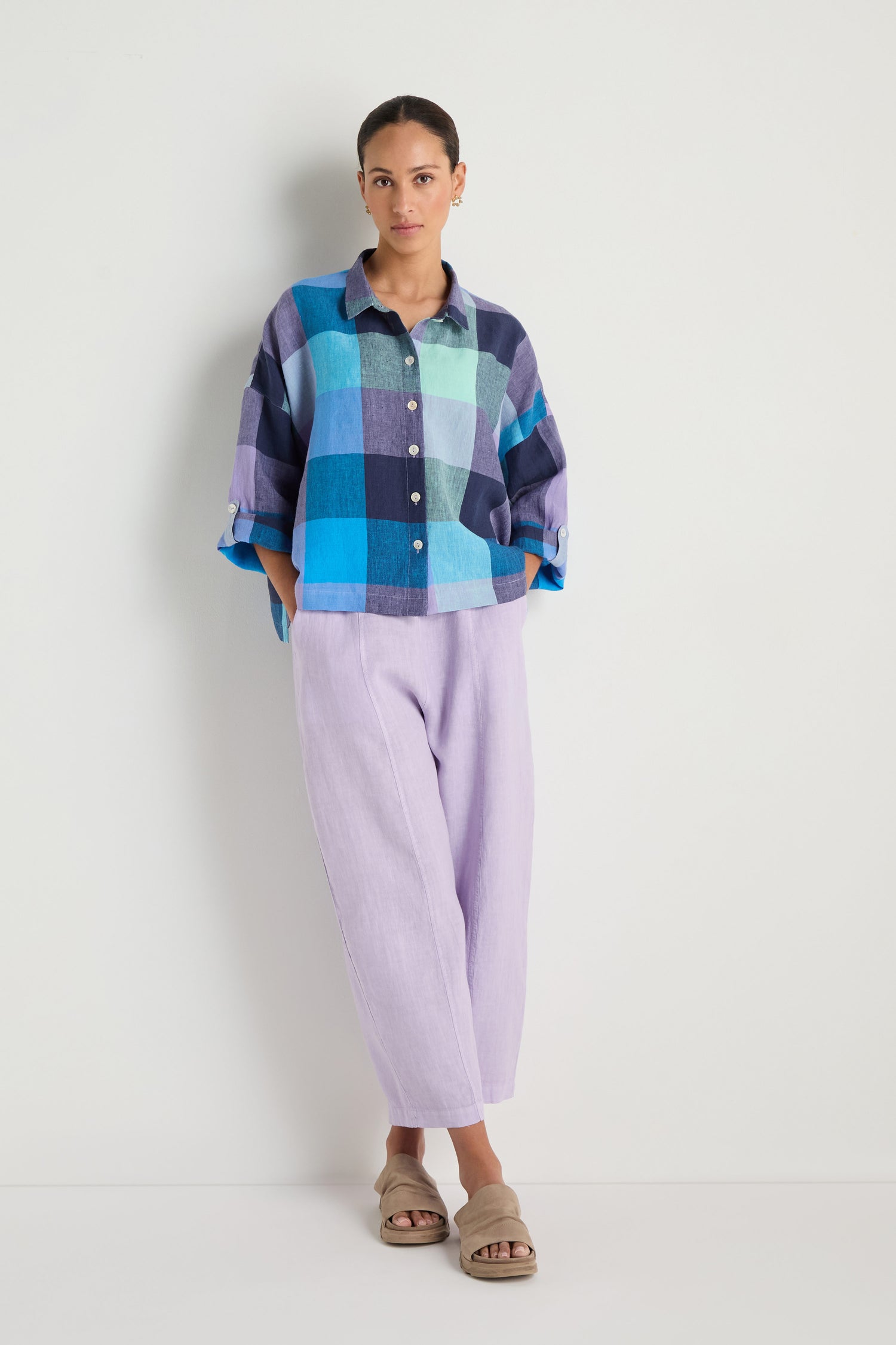 A woman with hands in pockets and a neutral expression stands against a white wall wearing an oversized fit Cross Dye Check Linen Shirt, light purple pants, and beige sandals.