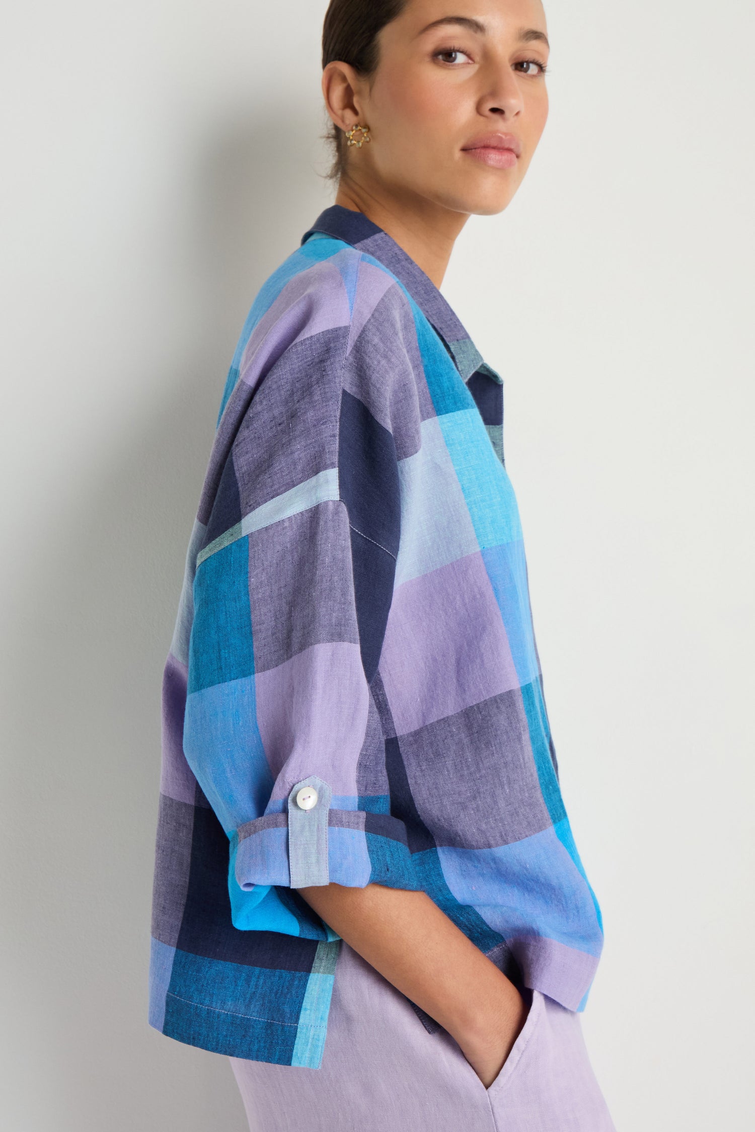 A person wearing the Cross Dye Check Linen Shirt, featuring an oversized blue and purple yarn-dyed check pattern and rolled sleeves, stands sideways with one hand in their pocket against a plain background.