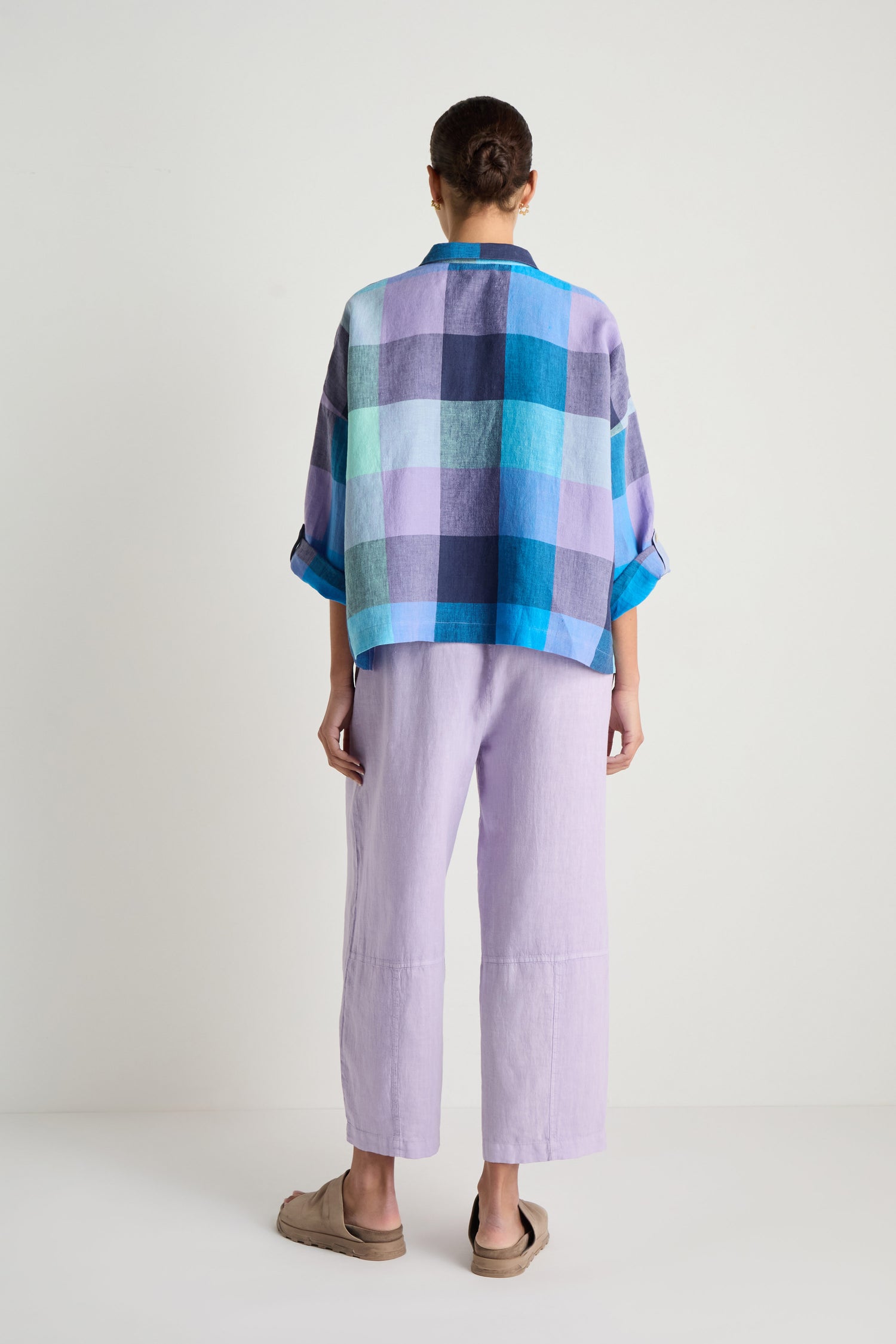 A person stands with their back to the camera, wearing a blue and purple Cross Dye Check Linen Shirt, light purple wide-leg oversized pants, and beige slip-on shoes against a plain white background.