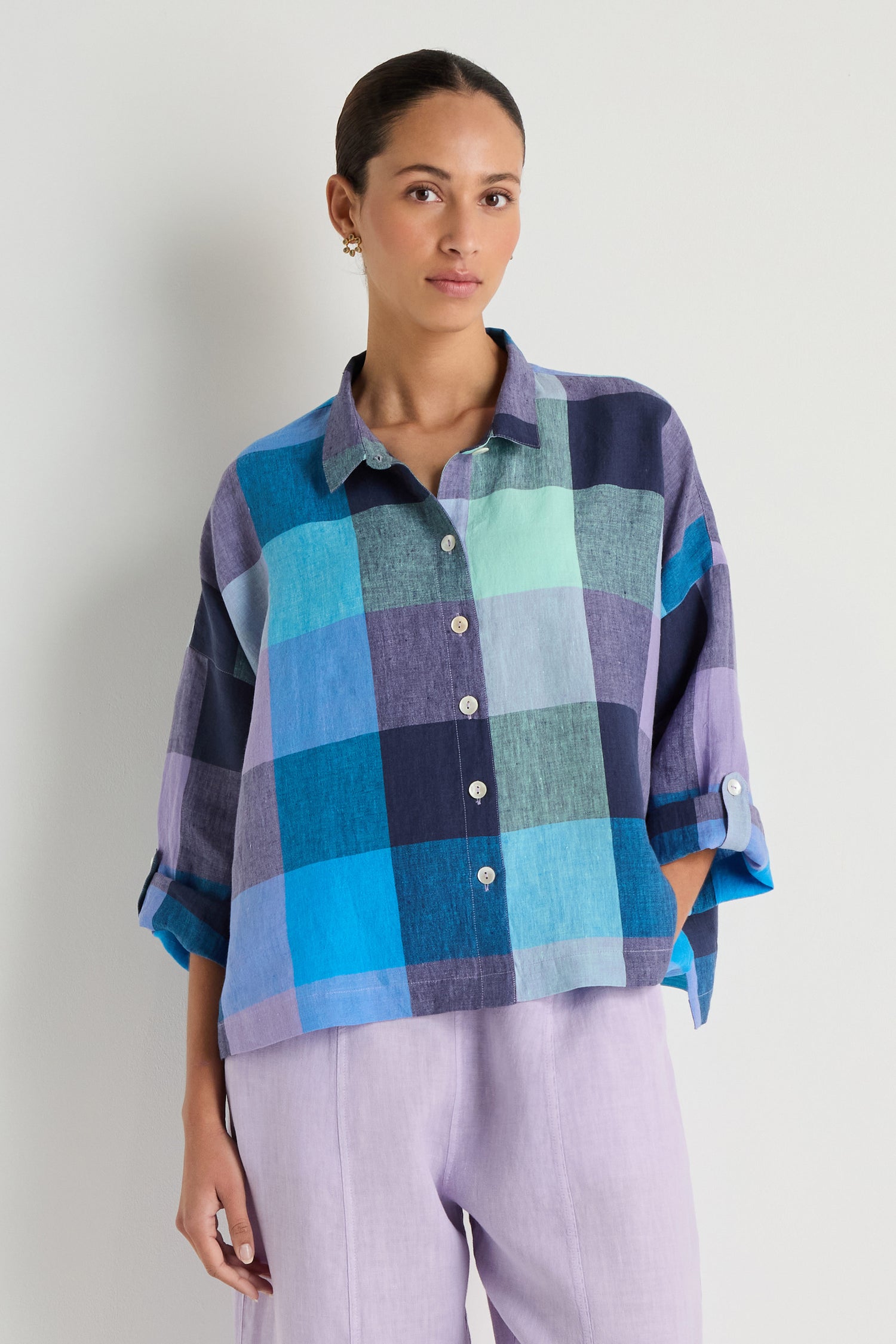 A woman stands against a plain background, wearing an oversized Cross Dye Check Linen Shirt with large blue and purple plaid checks and light lavender pants.