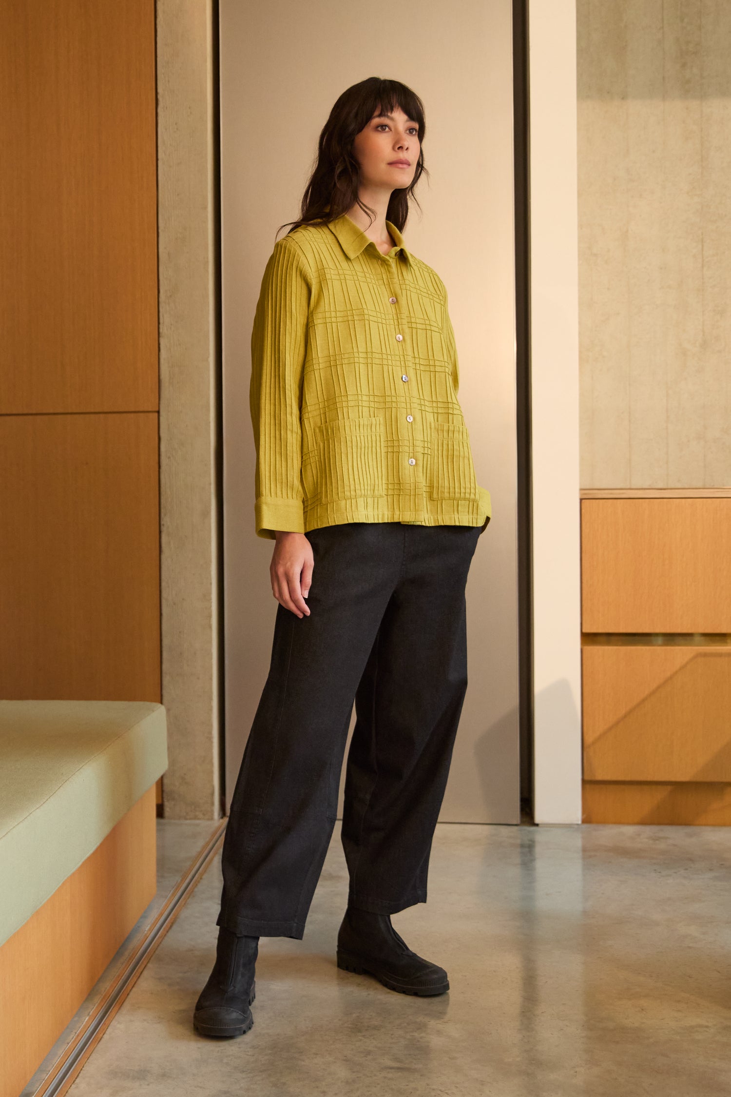 A woman stands indoors wearing the Brushed Cotton Pintuck Detail Shirt in yellow, paired with loose black pants and black shoes, with neutral-toned walls and furniture in the background.
