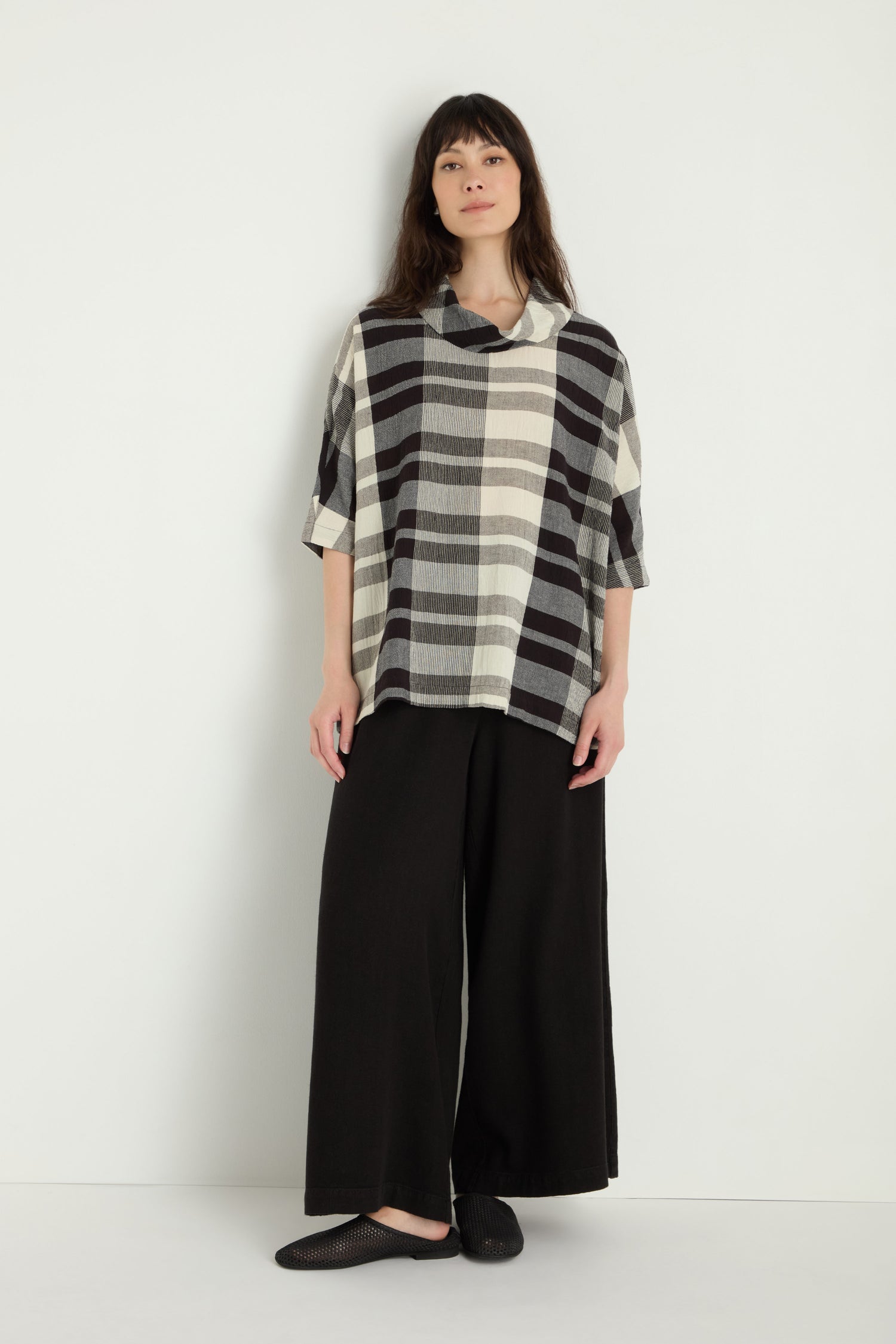 A woman stands against a white wall wearing the Monochrome Crinkle Check Cowl Neck Top, paired with wide-leg black pants and black shoes.