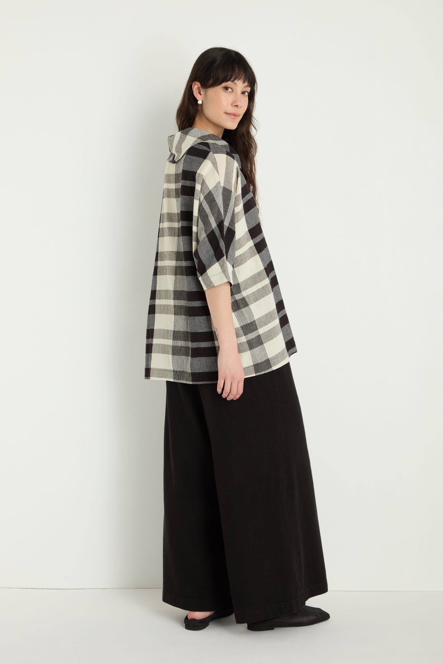 A woman stands against a white background, wearing the Monochrome Crinkle Check Cowl Neck Top with elbow-length sleeves and black wide-leg pants.